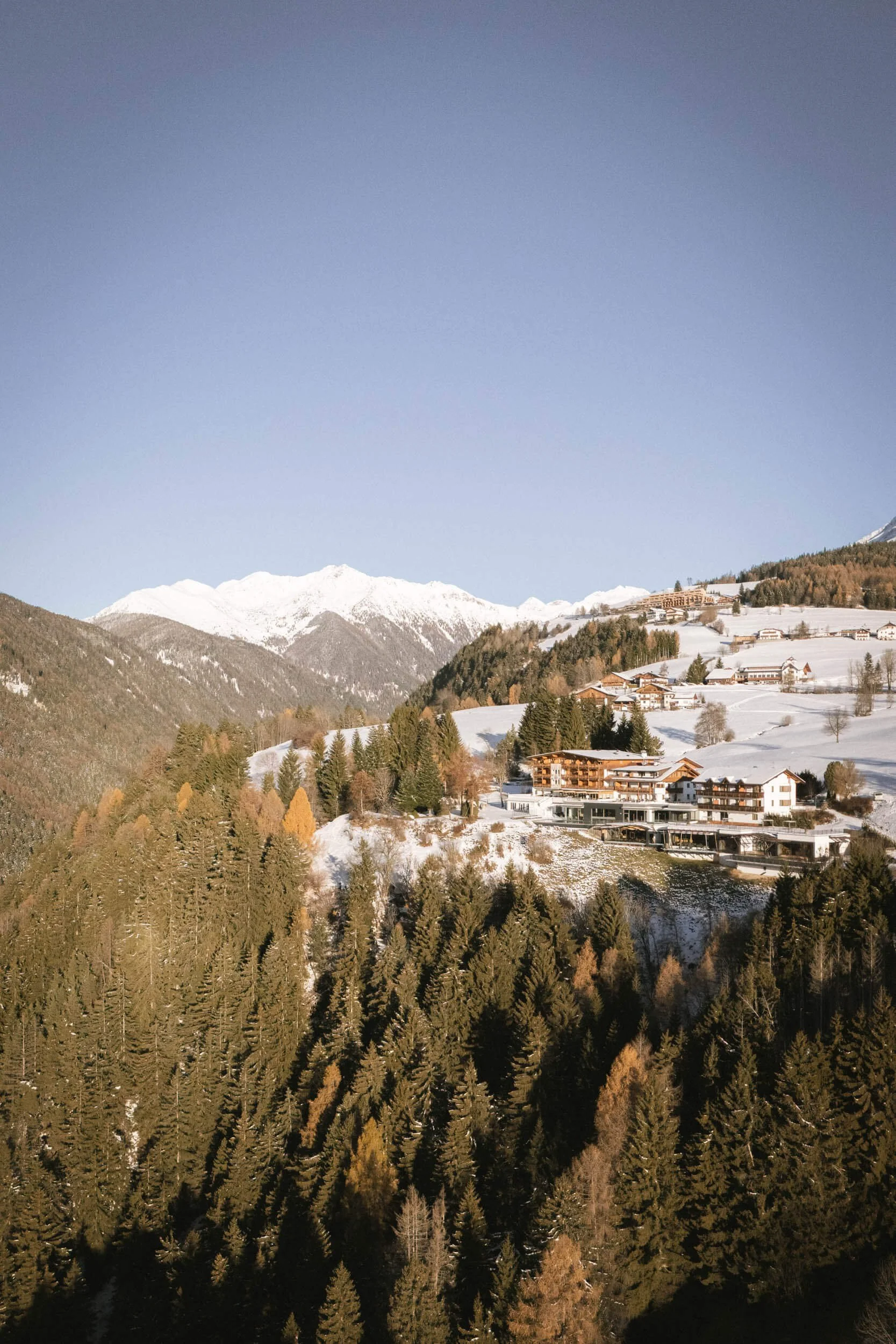 Schneebedeckte Berge, Häuser auf einer Hügellandschaft und Tannenbäume unter klarem Himmel.