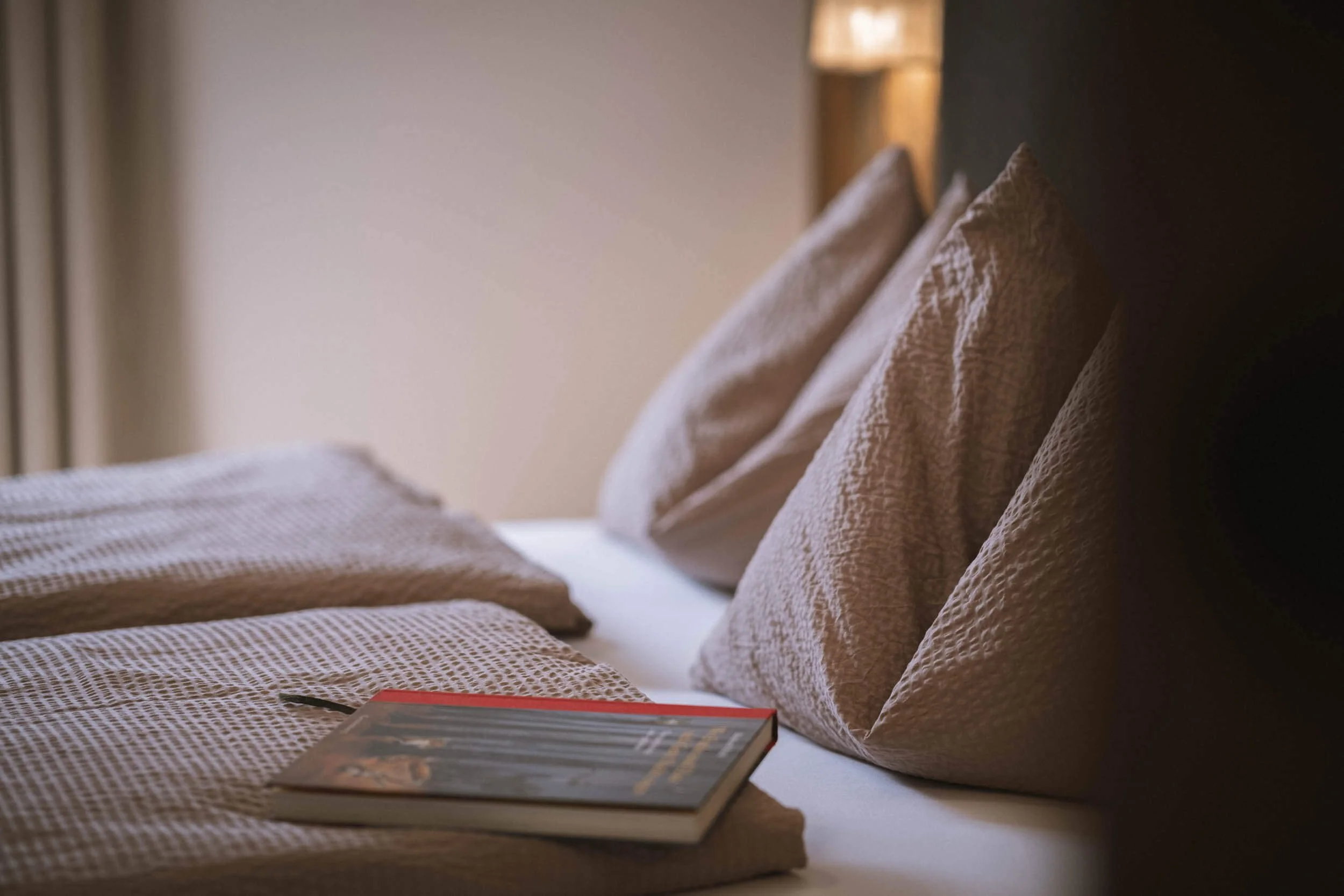 Bett mit weißen Kissen, Bettdecke und einem aufgeschlagenen Buch mit einem Stift darauf.