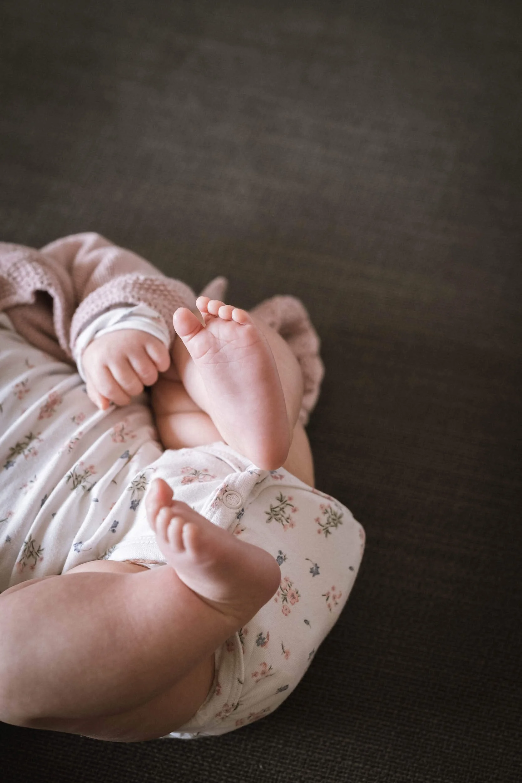 Nahaufnahme eines Babys, das auf einem dunklen Boden liegt, mit einem Arm, der gehalten wird, und nackten Füßen, die in die Luft zeigen.