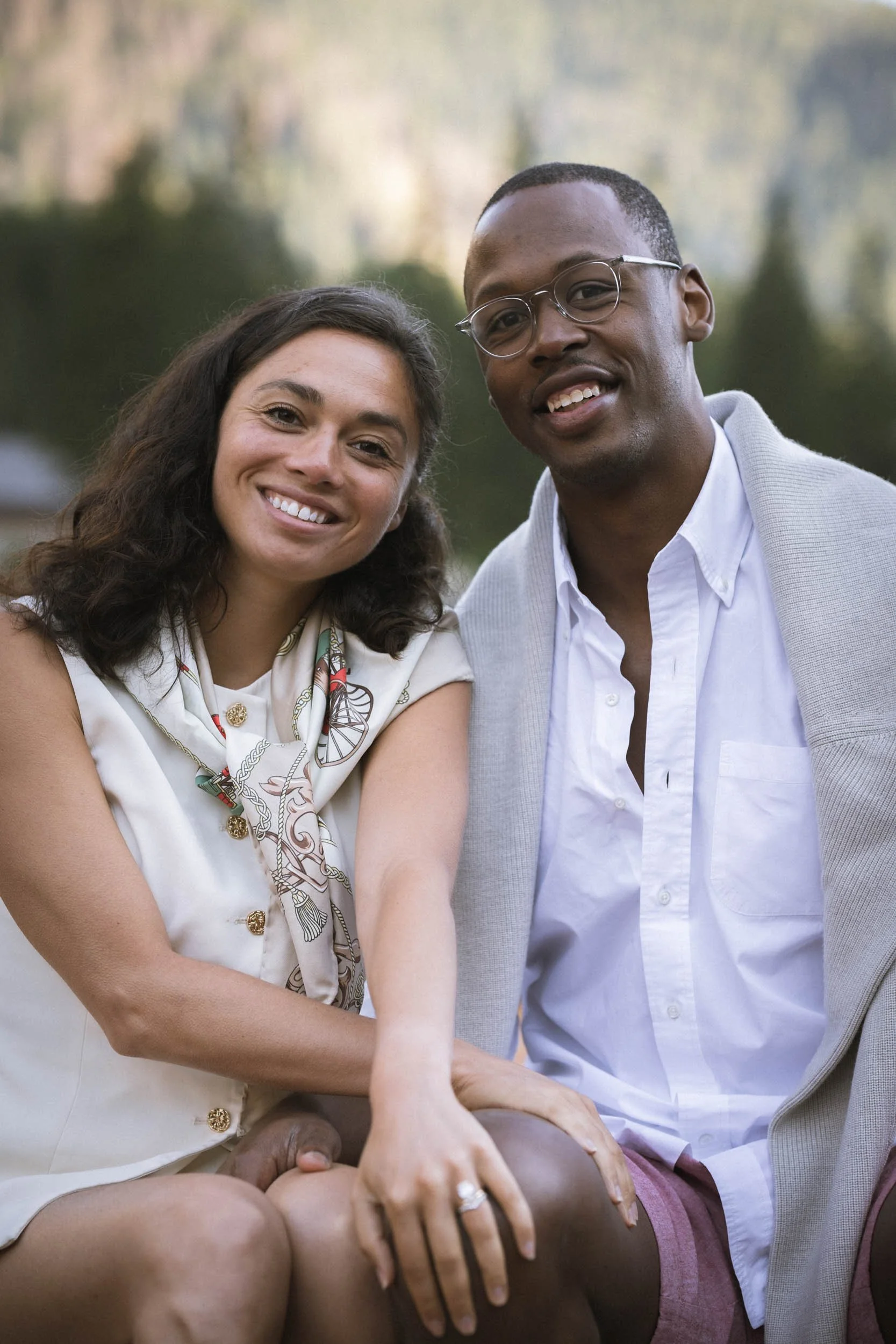 Ein lachendes Paar, das im Freien sitzt, umgeben von Natur, mit der Frau, die die Hand ihres Partners hält.