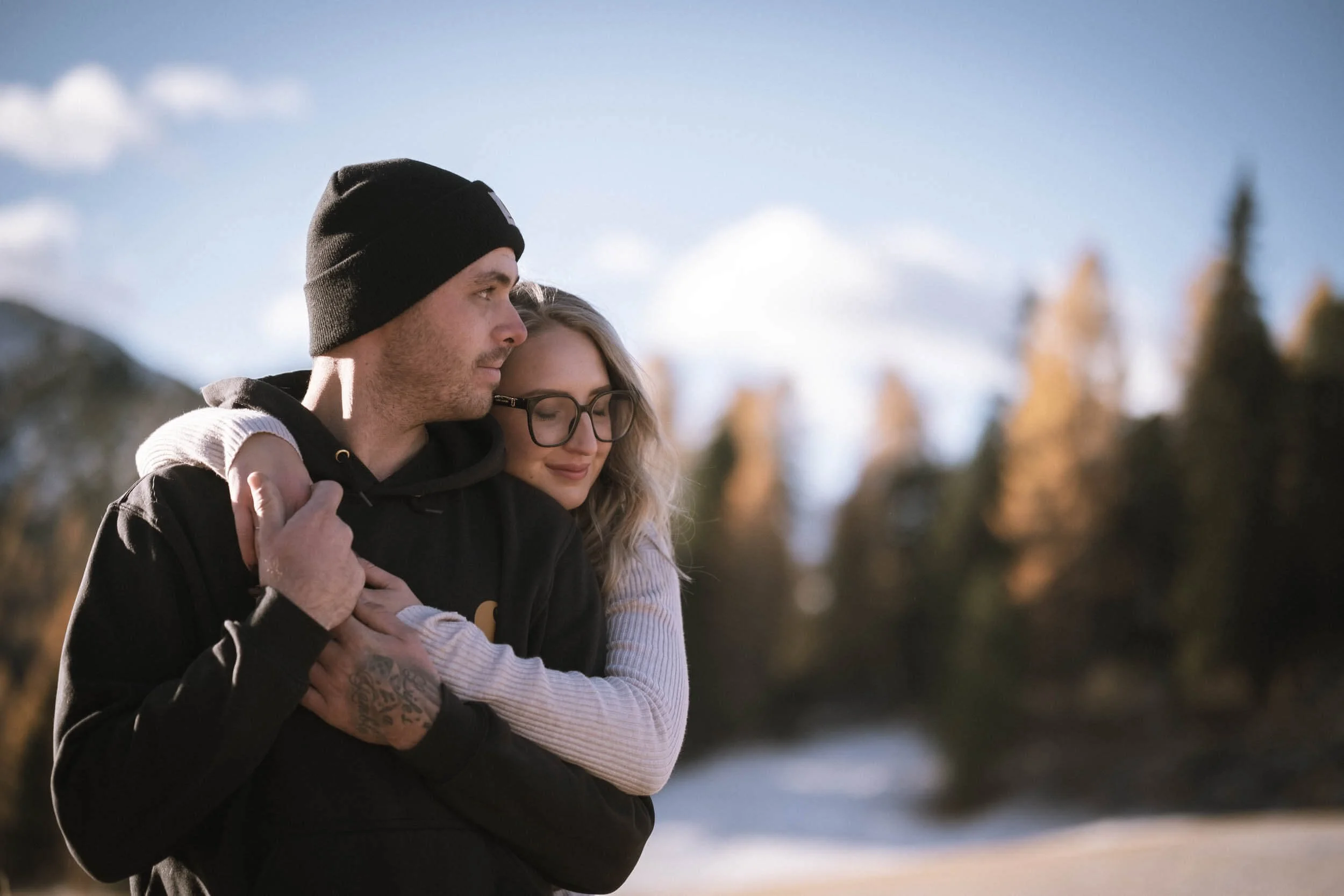 Ein Mann und eine Frau umarmen sich draußen bei schönem Wetter, im Hintergrund sind Bäume und Himmel.