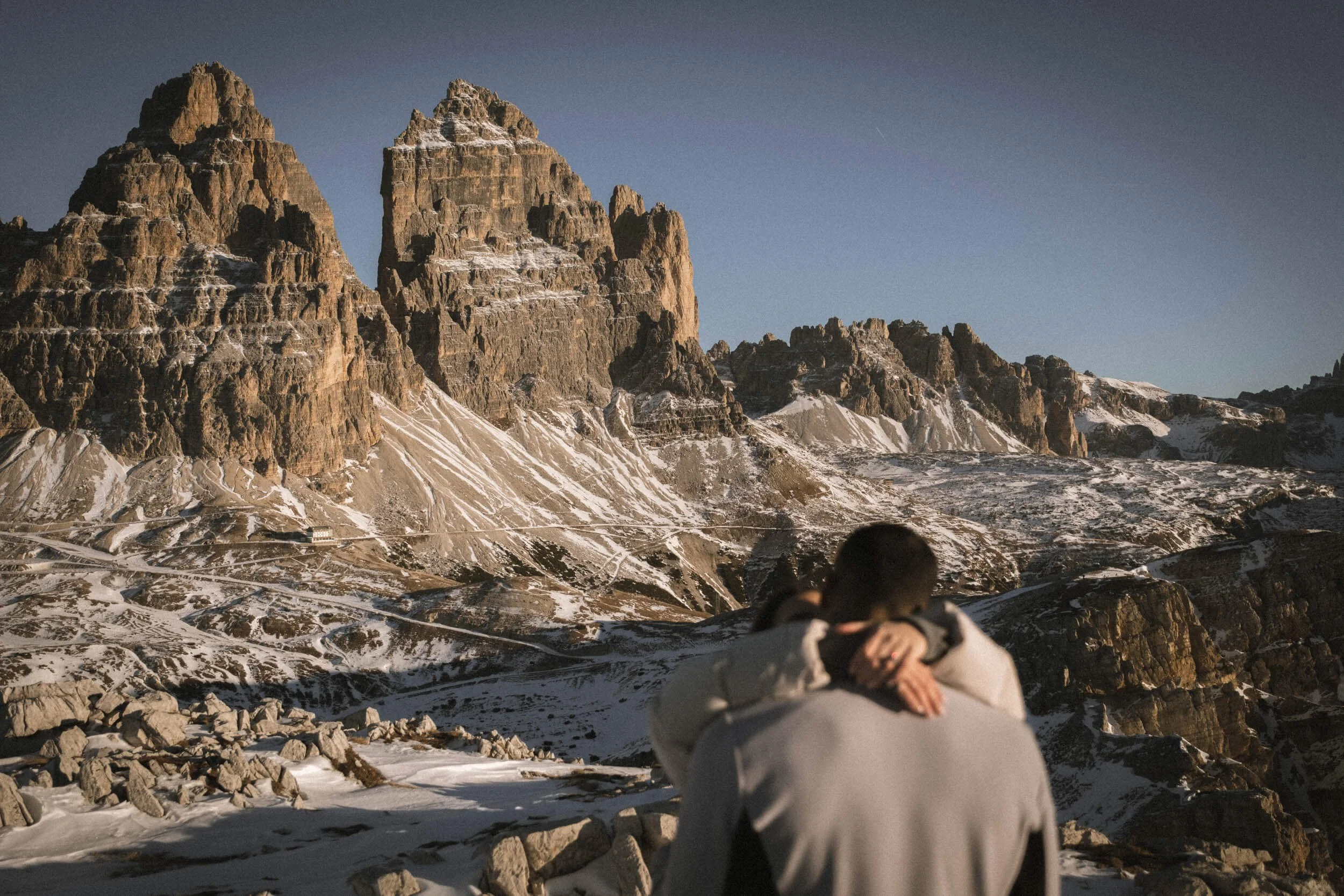 Zwei Personen in einem verschneiten Gebirge, die sich umarmen, mit dramatischen Felsformationen im Hintergrund.