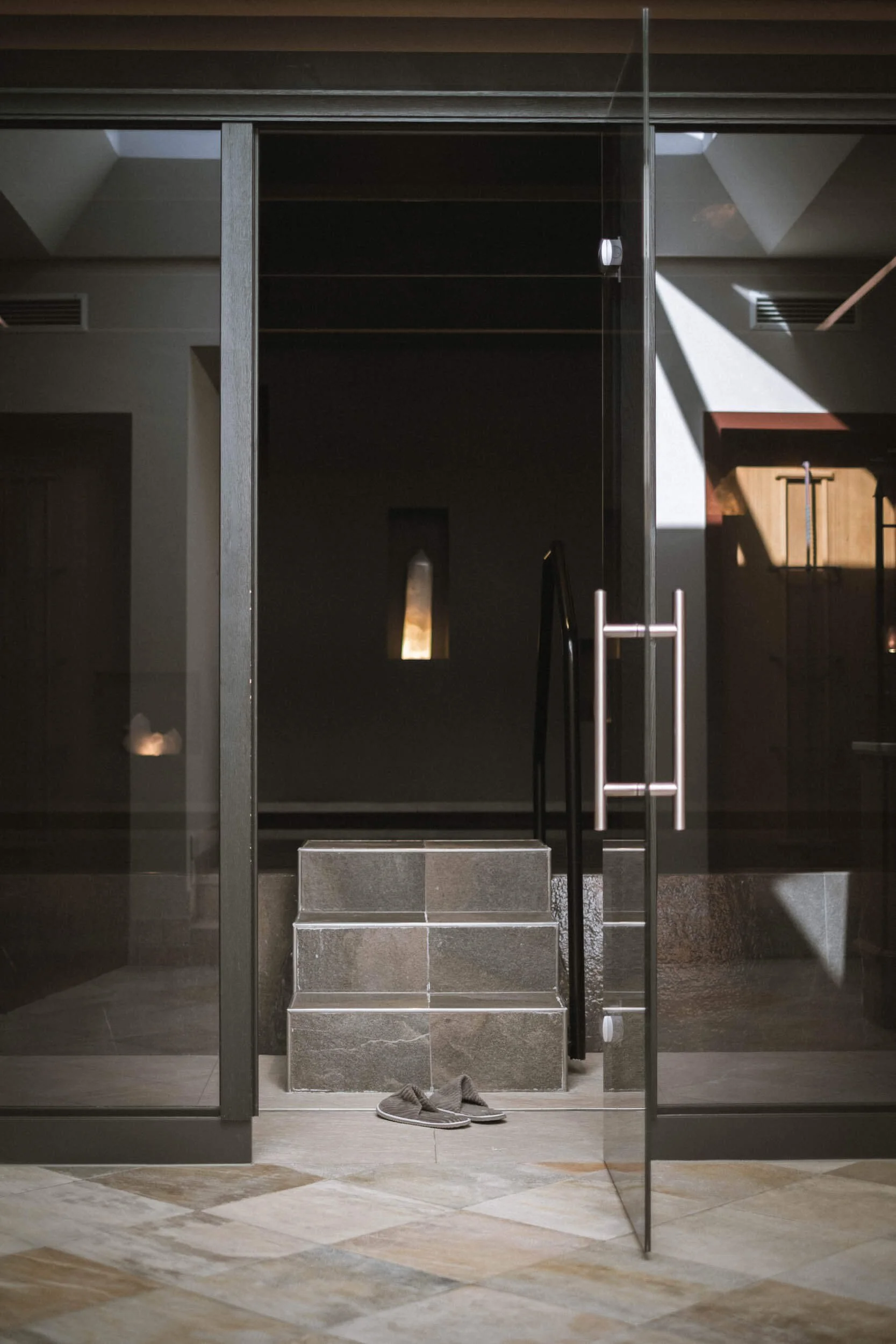 Ein dunkler Eingangsbereich mit Stufen aus Stein, darüber ein Glasdach und eine offene Tür, Schuhe auf dem Boden, Sonnenlicht fällt durch ein Fenster.