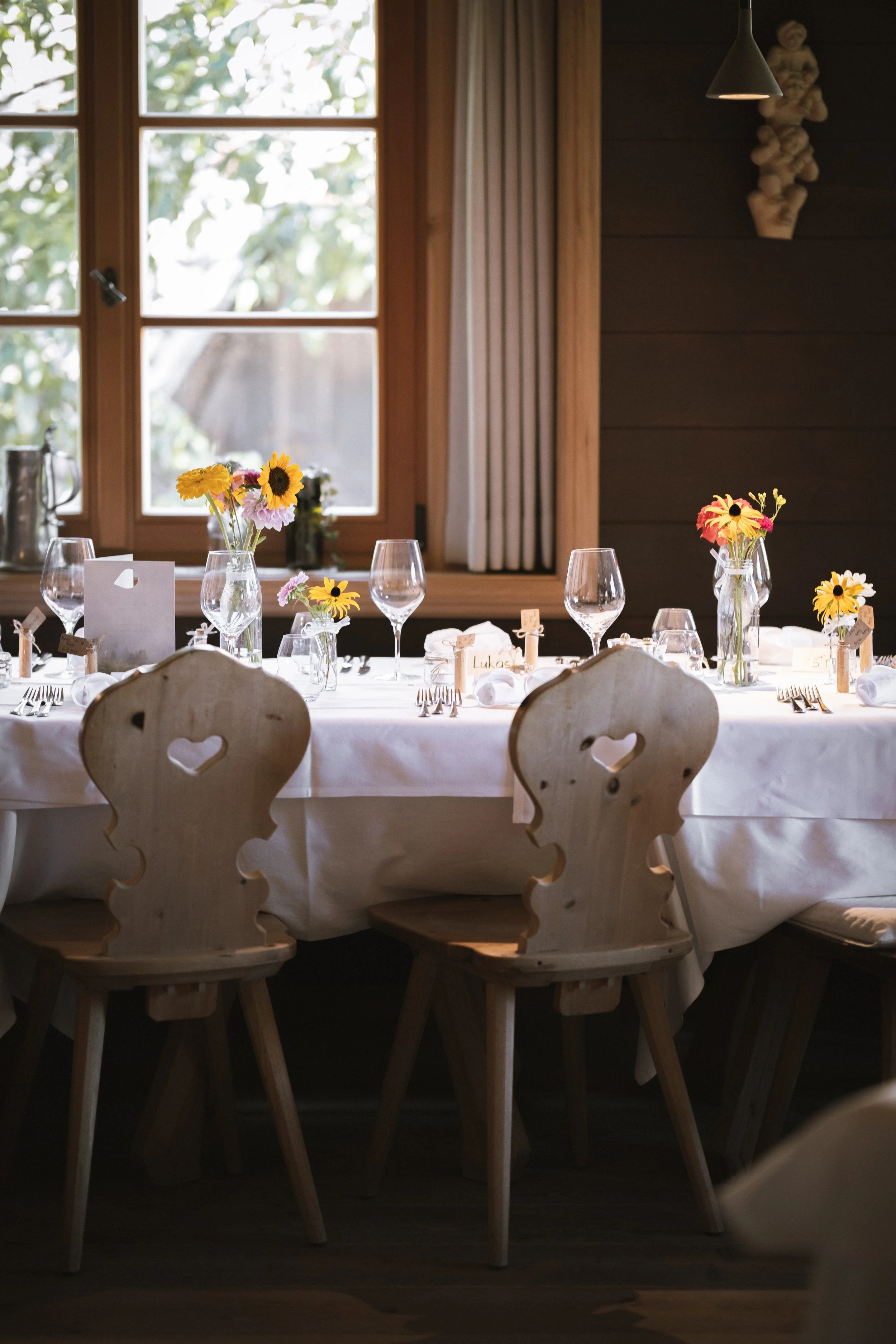 Ein festlich gedeckter Tisch mit Blumen, Weingläsern und Namenetiketten in einem rustikalen Raum mit Holzvertäfelung und Fenster. 