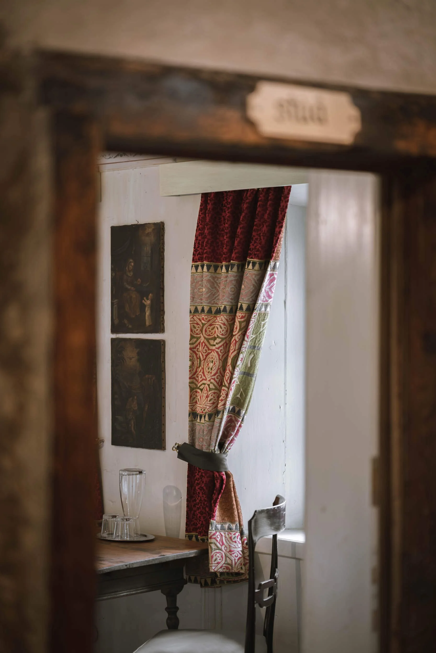 Ein Blick durch ein geöffnetes Türrahmen zeigt einen Raum mit einer Holztisch, zwei Gläsern, einem weißen Gefäß, einem Stuhl und einem Fenster mit schweren, bunten Vorhängen. An der Wand hängen zwei alte, dunkle Gemälde.