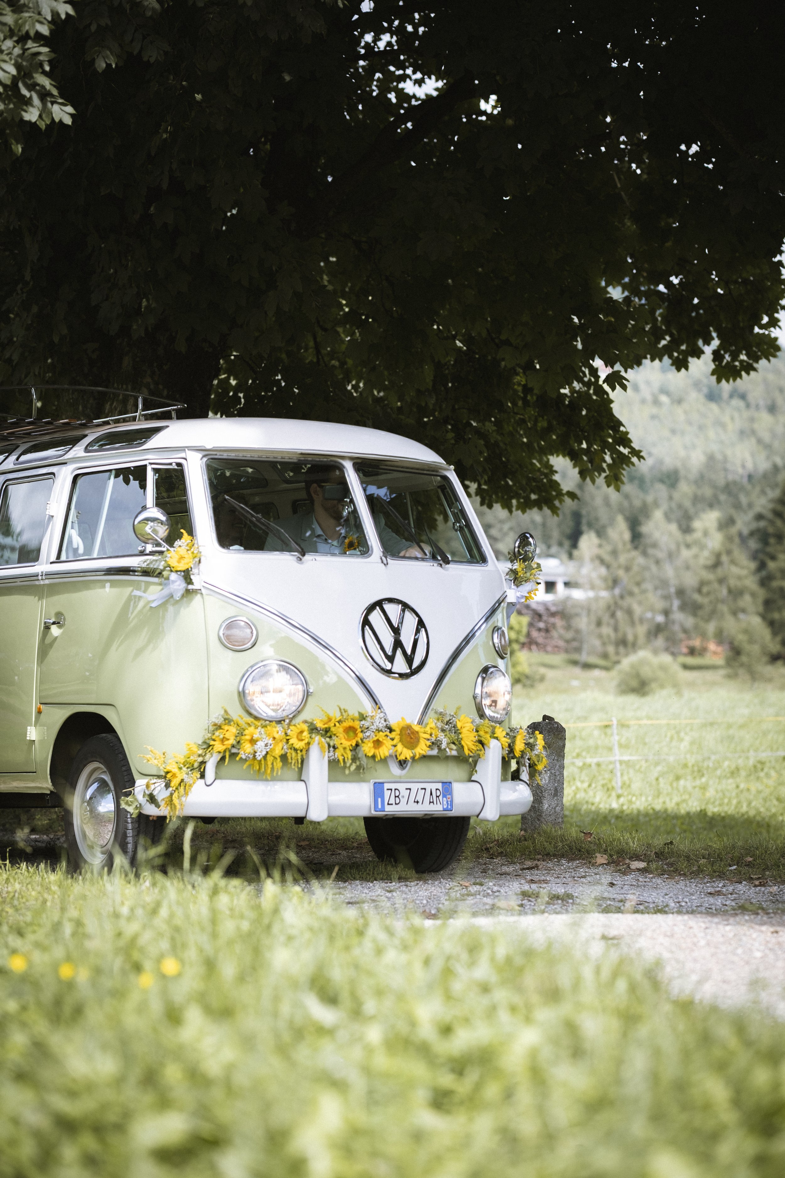 Ein grün-weißer VW-Bulli, verziert mit Sonnenblumen, steht auf einem Feld unter einem Baum, möglicherweise für eine Hochzeit oder eine Feier.