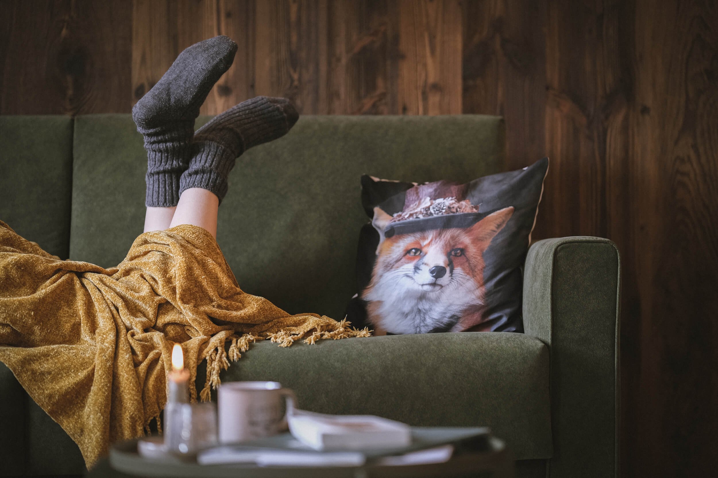 Eine Person liegt auf einem grünen Sofa, trägt schwarze Stricksocken, und hat eine braune Decke über den Beinen. Auf dem Sofa liegt ein Kissen mit einem Fuchsgesicht. Im Vordergrund steht eine Tasse auf einem Tisch, und eine Kerze ist ebenfalls sicht