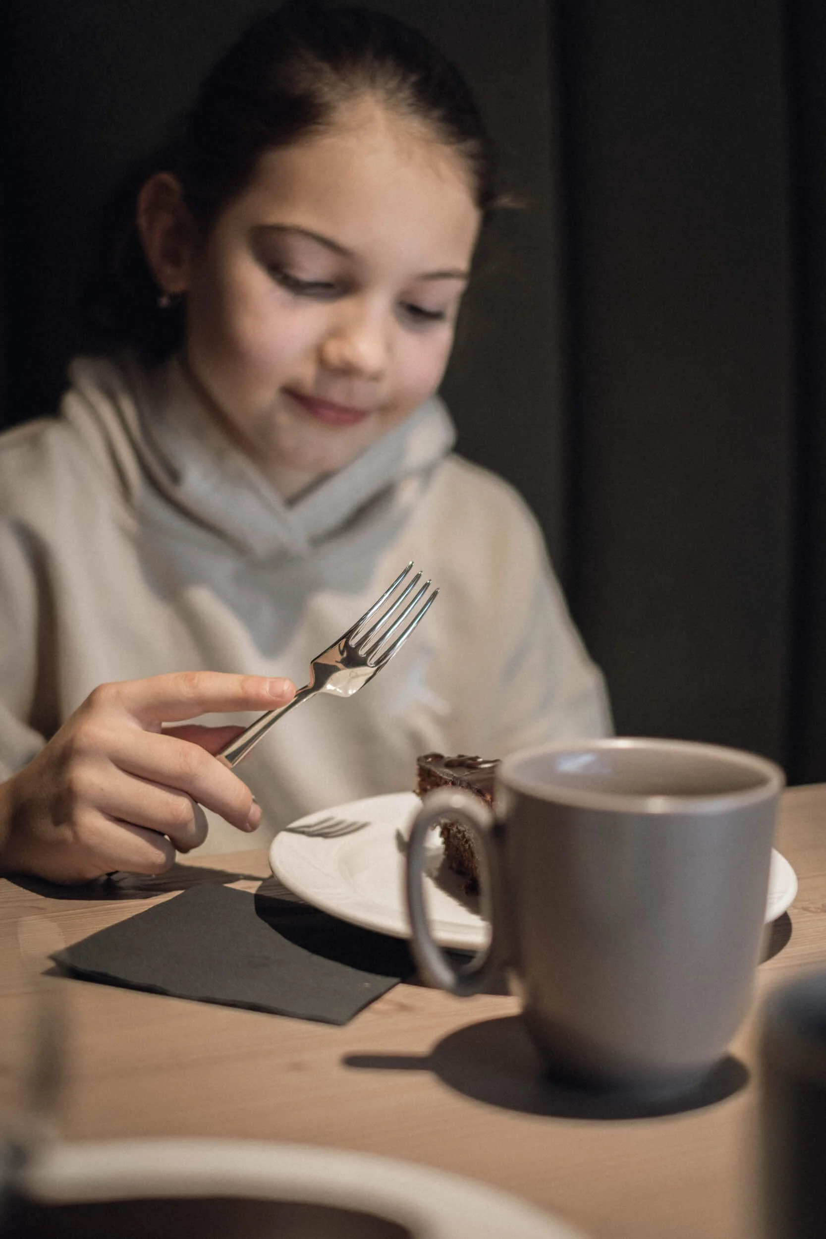 Junge Frau isst ein Stück Schokoladentorte mit einer Gabel, auf einem Teller, neben einer weißen Tasse. Sie sitzt an einem Holztisch in einem dunklen Raum.