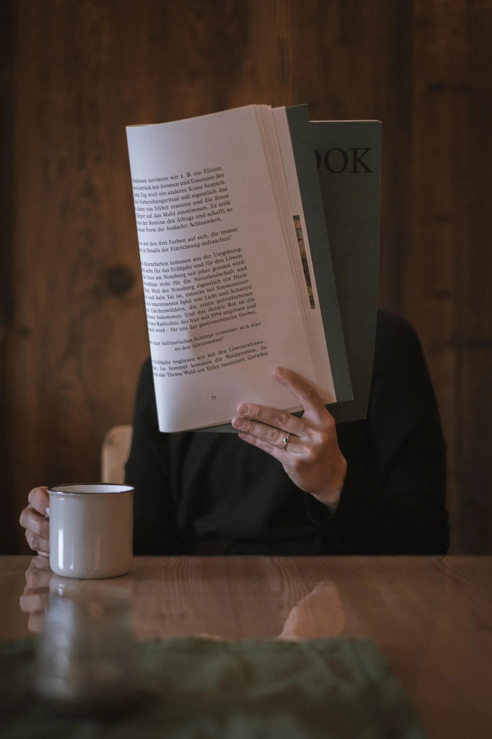 Person liest eine Zeitschrift an einem Holztisch, hält eine Tasse in der anderen Hand, im Hintergrund eine dunkle Holzwand.