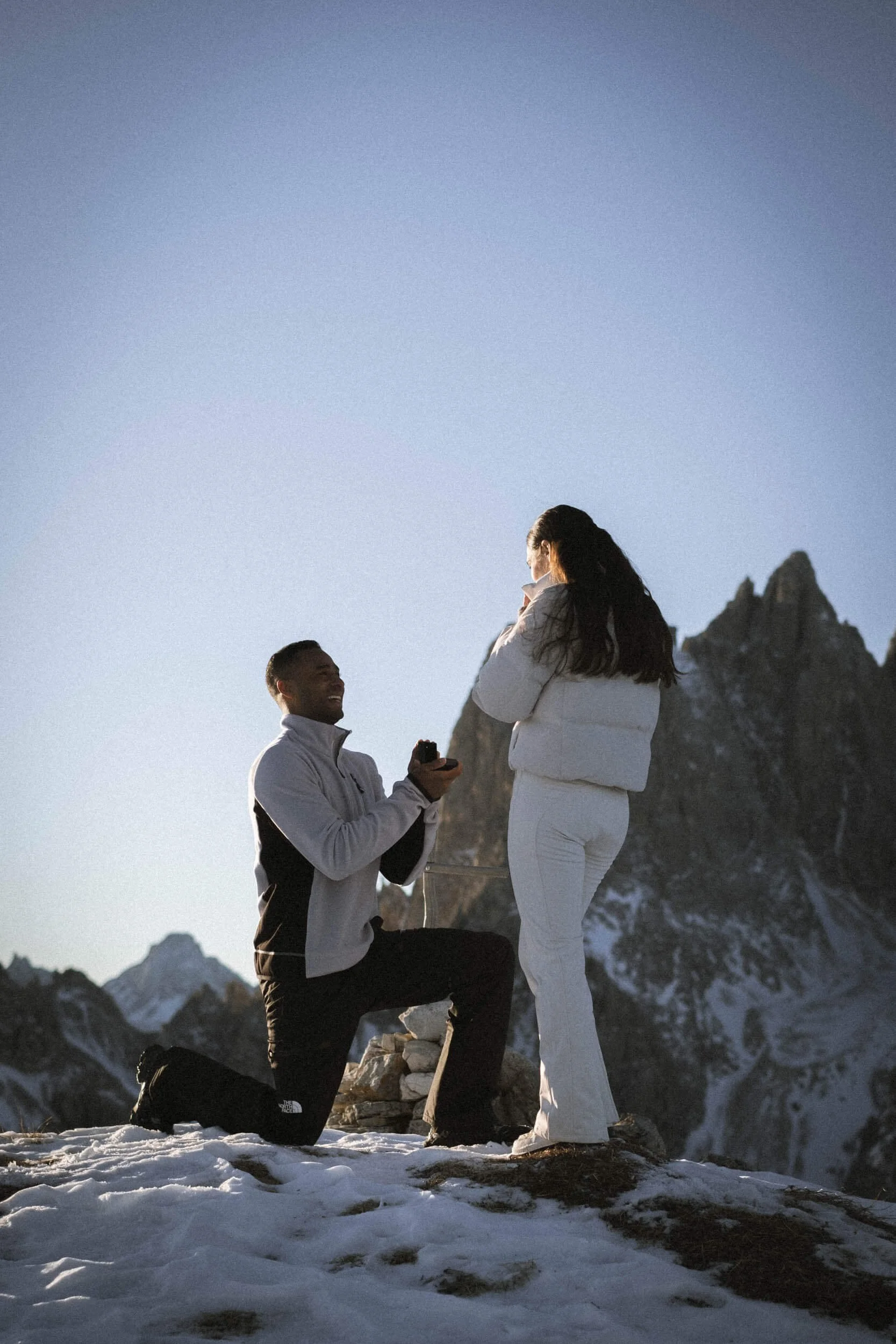 Verlobung auf einem schneebedeckten Berggipfel bei Sonnenaufgang, Mann kniet mit einem Verlobungsring, Frau überrascht