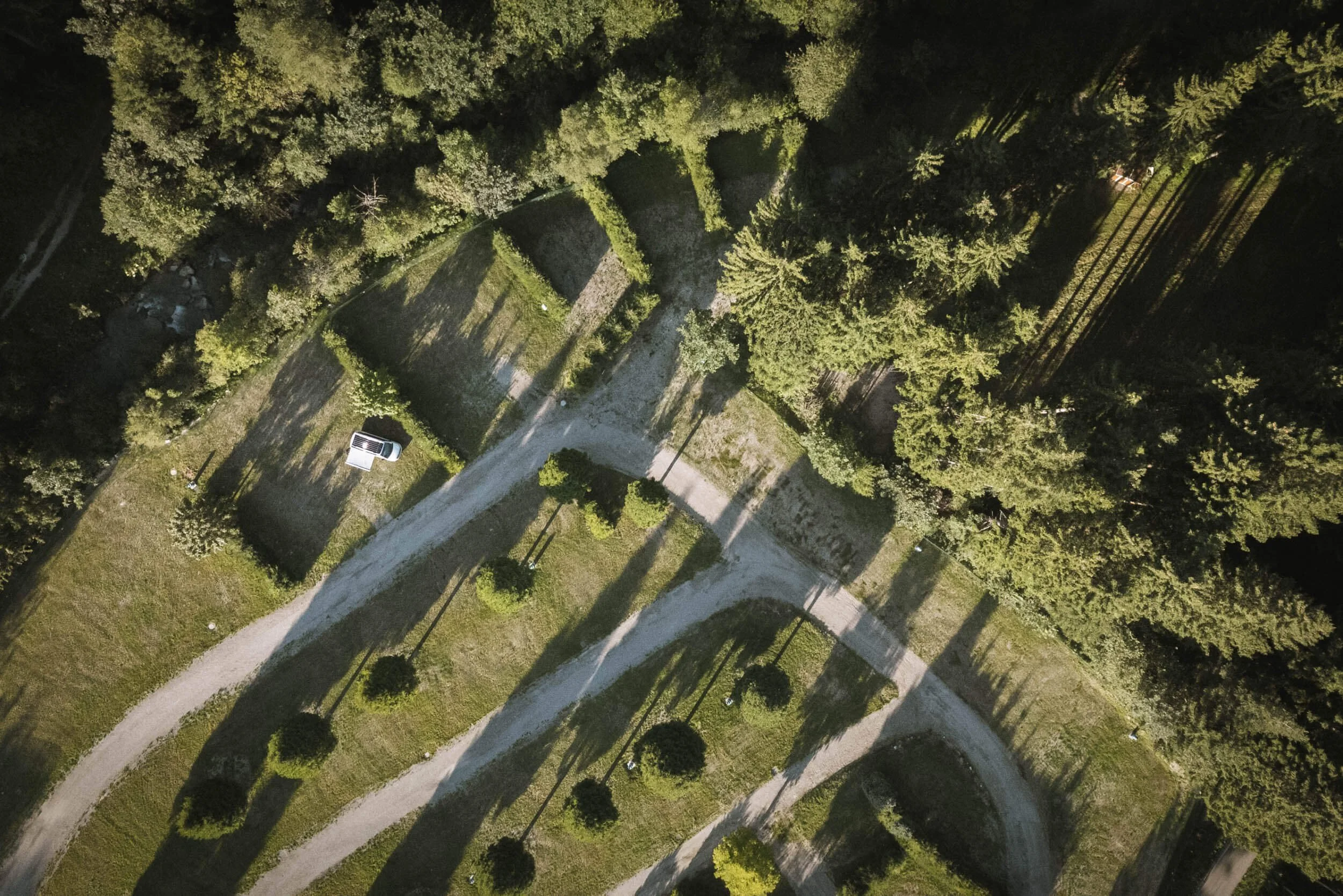 Luftaufnahme eines Gartens mit Wegen, Bäumen und Schatten, sowie einem weißen Fahrzeug auf dem Rasen.