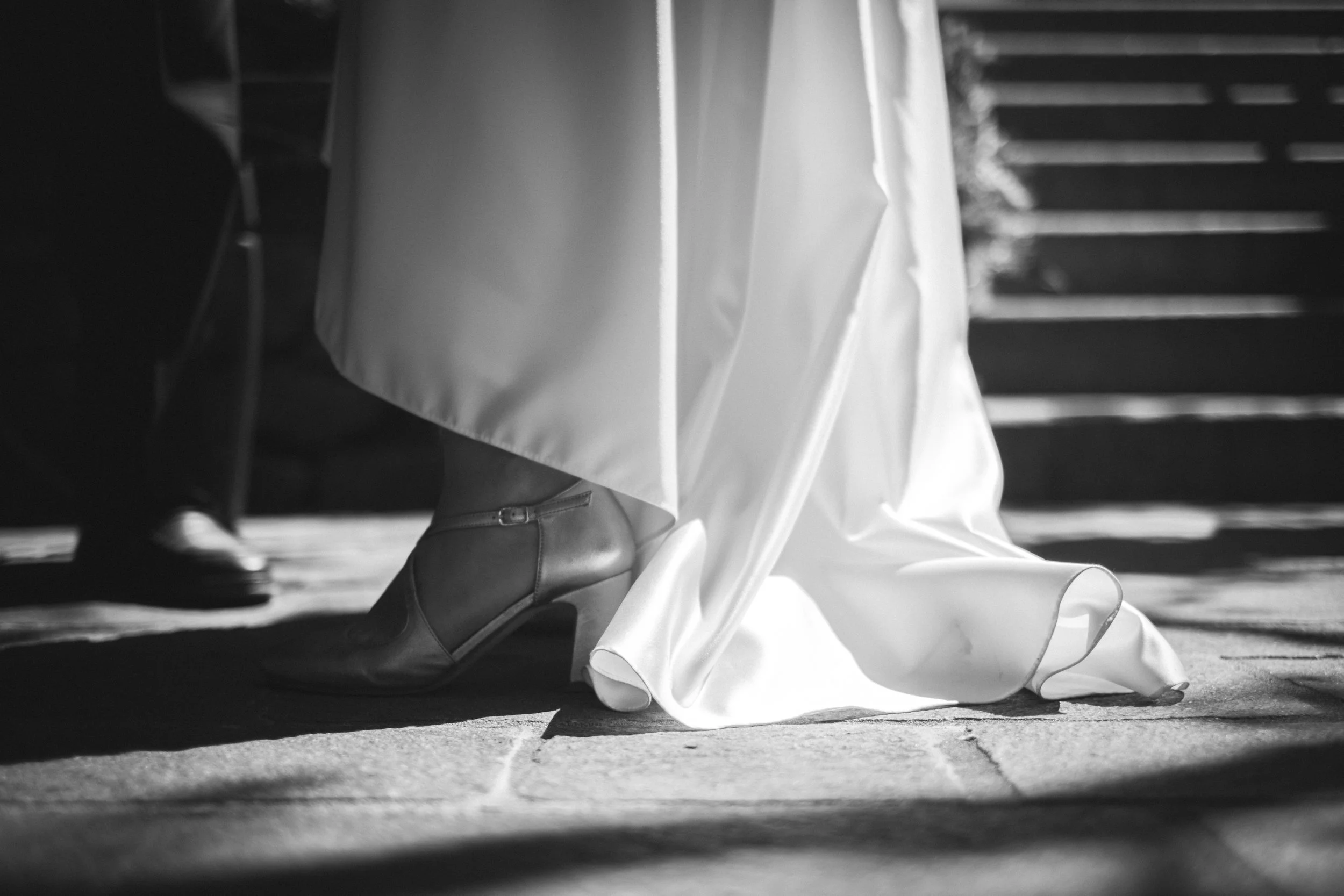 Schwarz-Weiß-Foto eines Brautpaares, bei dem der Fokus auf den Schuhen der Braut liegt, die unter einem langen weißen Kleid hervorlugen.