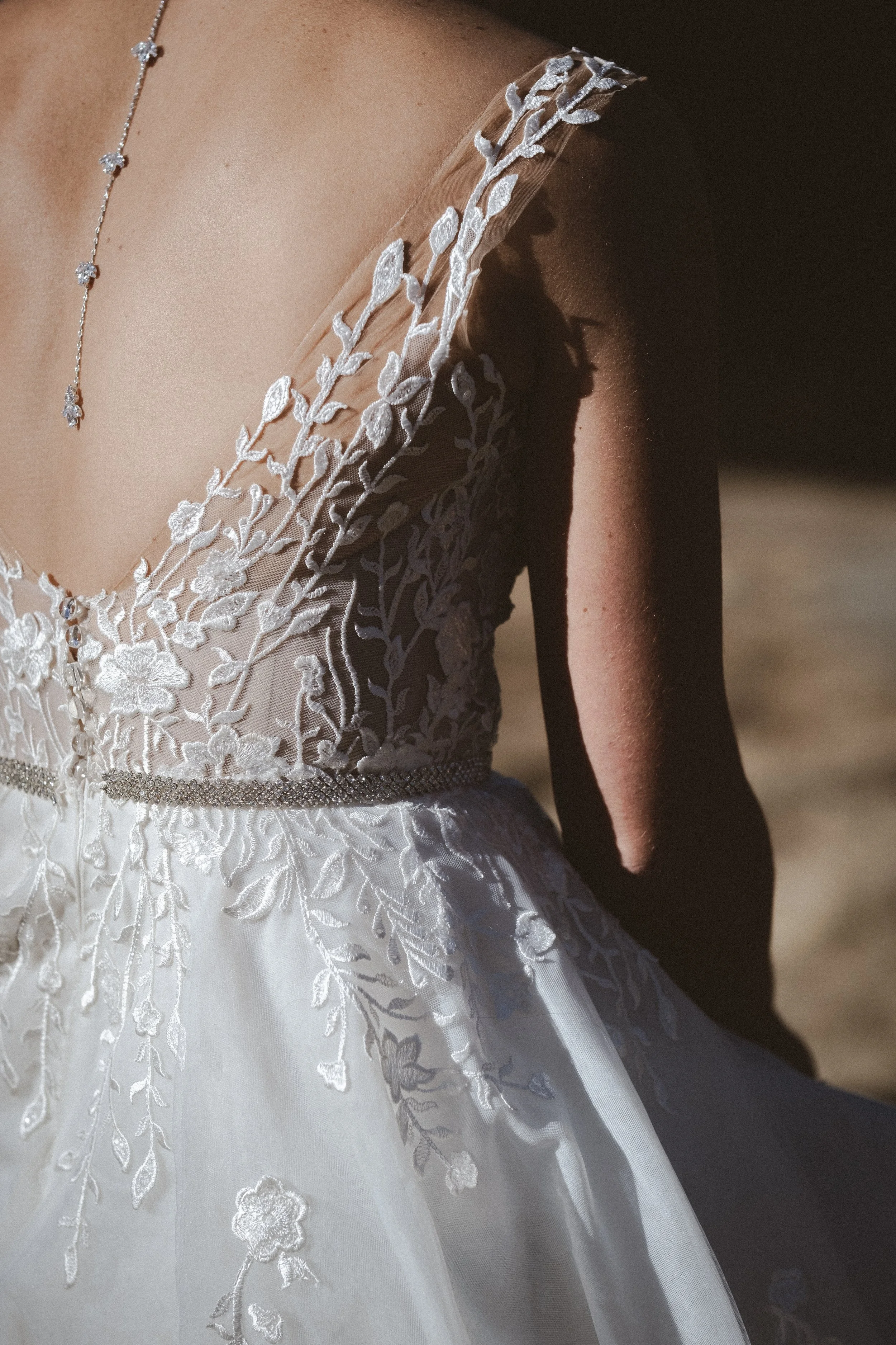 Nahaufnahme eines Brautkleides mit aufwendiger Stickerei und transparenten Trägern, getragen wird eine dezente Halskette.
