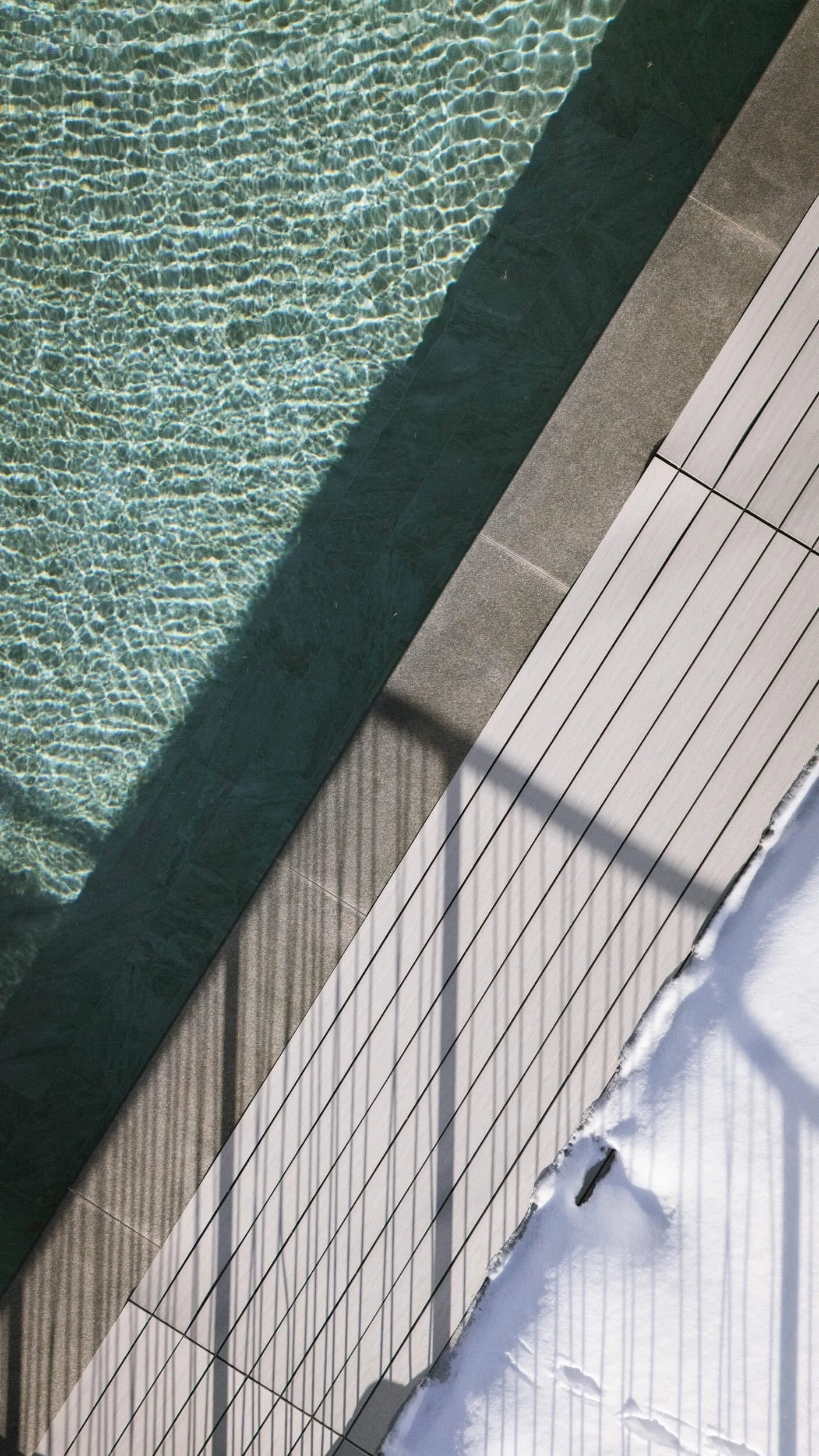 Blick auf einen Pool mit Wasser und eine Holzplattform mit Schatten auf Schnee, bei Sonnenlicht aufgenommen.