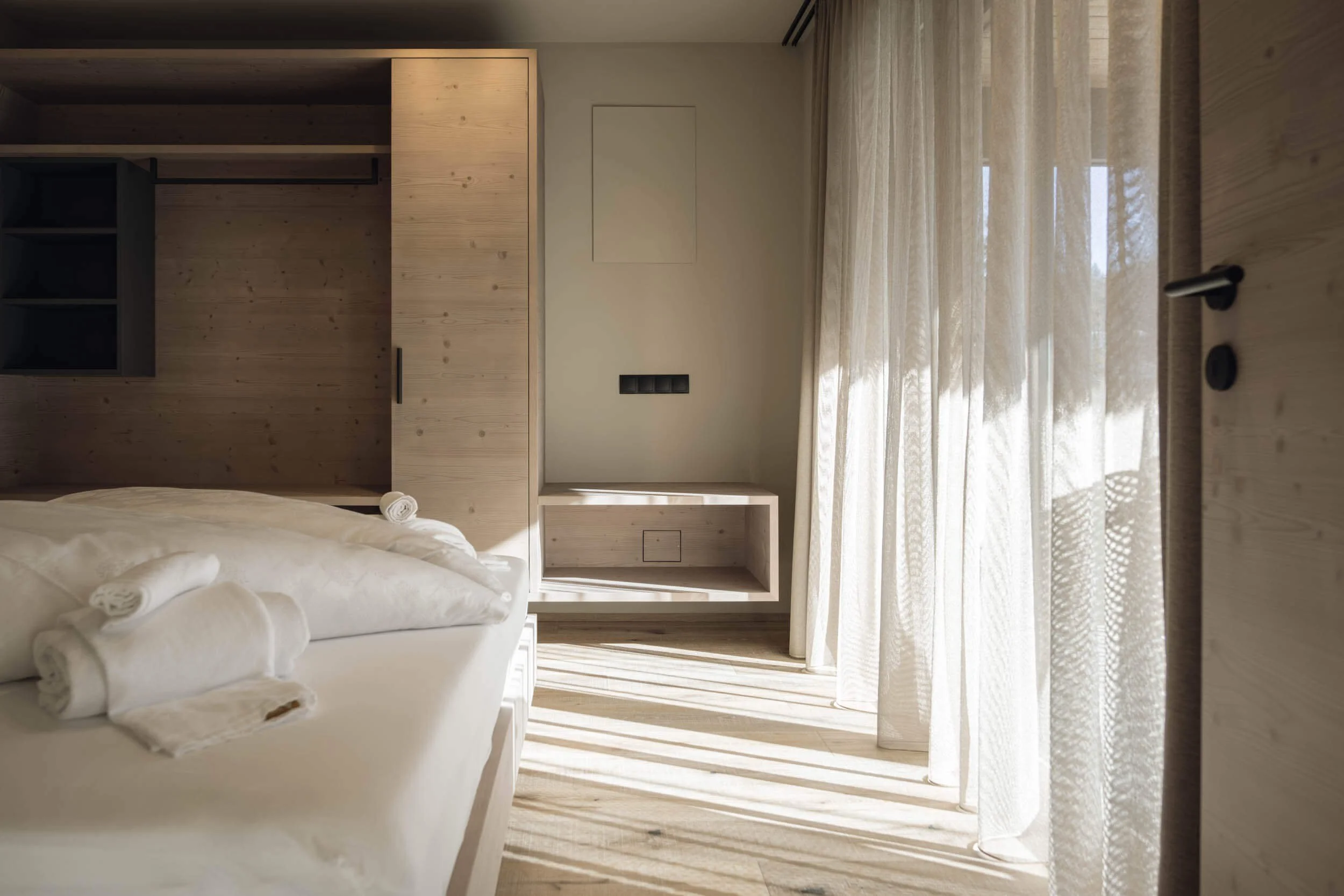 Ein modern eingerichtetes Hotelzimmer mit Holzdekoration, einem Bett mit weißen Handtüchern, großen Fenstern mit transparenten Gardinen, und einem natürlichen Farbthema.