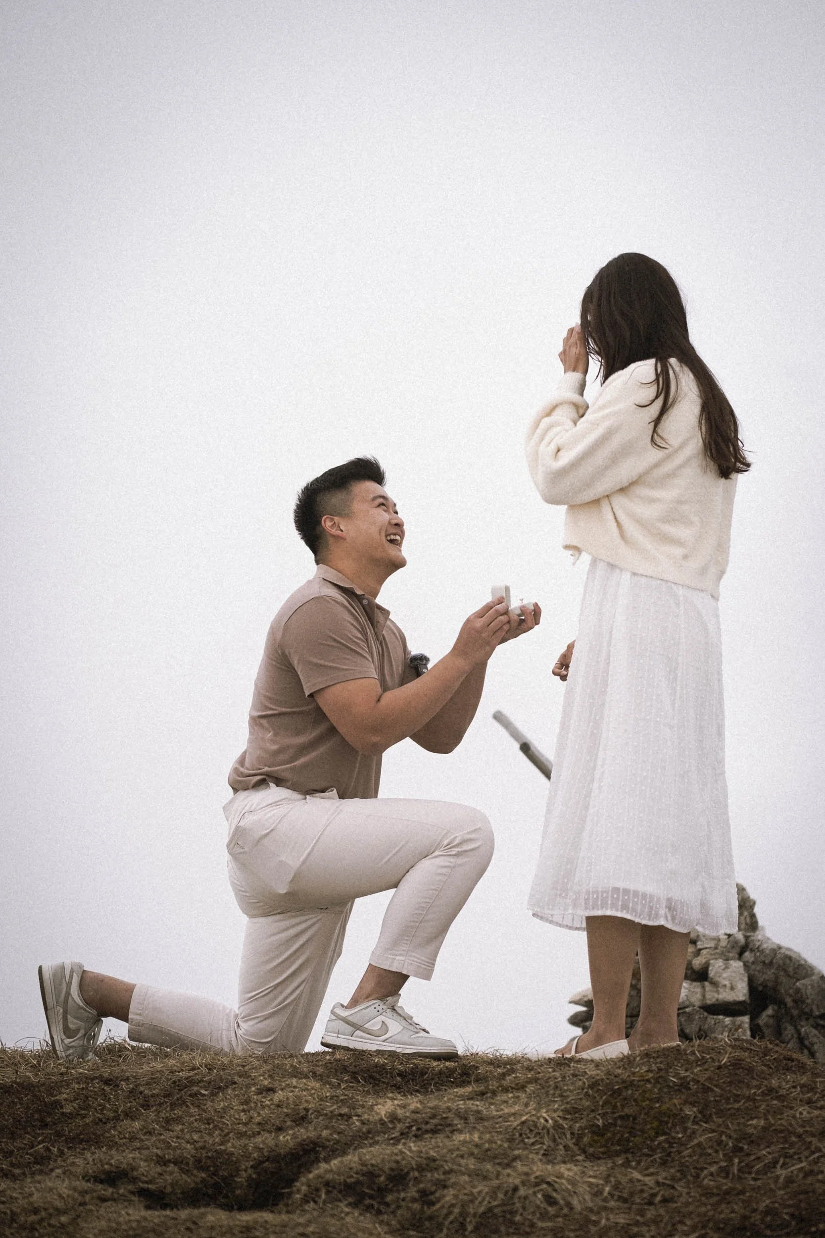 Ein Mann macht einem Mädchen einen Heiratsantrag auf einem Hügel im Freien, beide lachen glücklich.