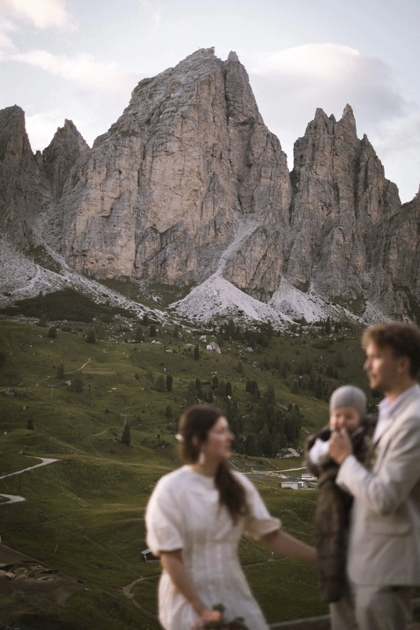 Ein Mann hält ein Kleinkind in den Armen, während eine Frau neben ihm steht, vor einer Berglandschaft mit hohen Felsen und grünen Wiesen.