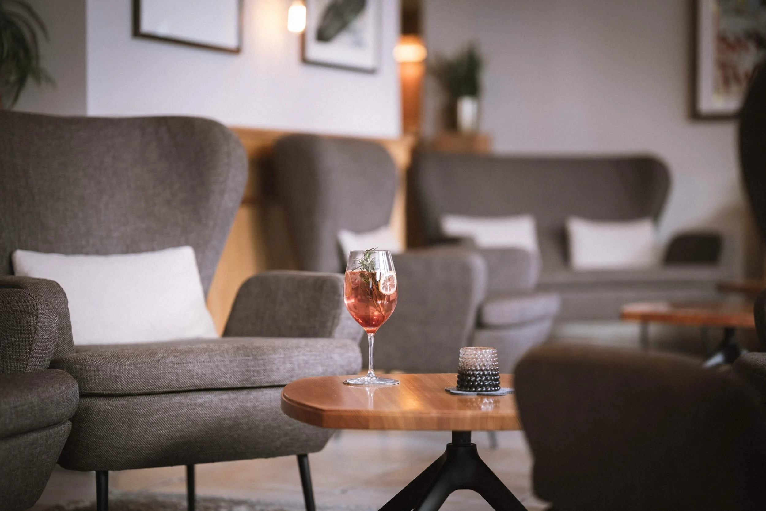 Ein Glas Roséwein mit Zitronenscheibe und Rosmarin auf einem runden Holztablett in einem Wohnzimmer mit grauen Sofas, weißen Kissen und Bilder an der Wand.