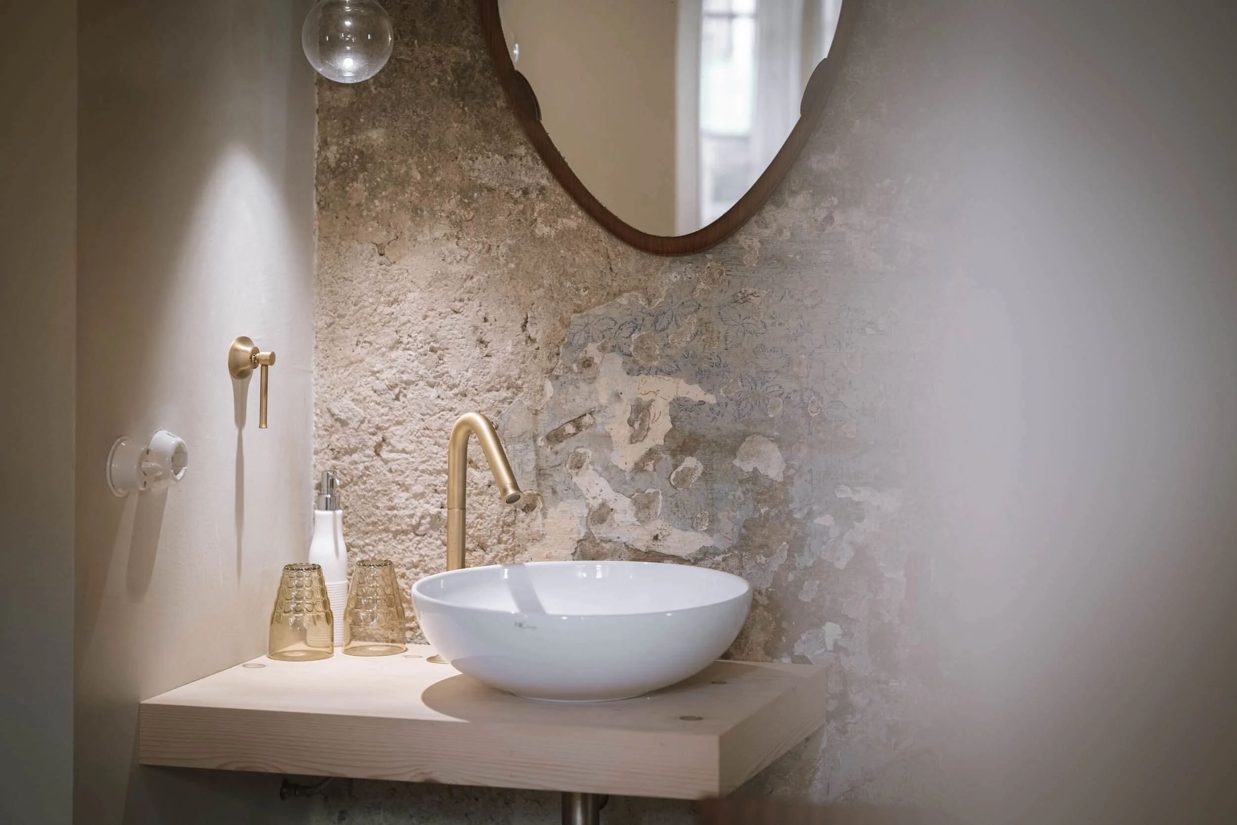 Ein Waschbecken mit goldener Armatur vor einer Wand mit einer Halbkugel Spiegel und einer Wand mit abblätternder, alter Putz.