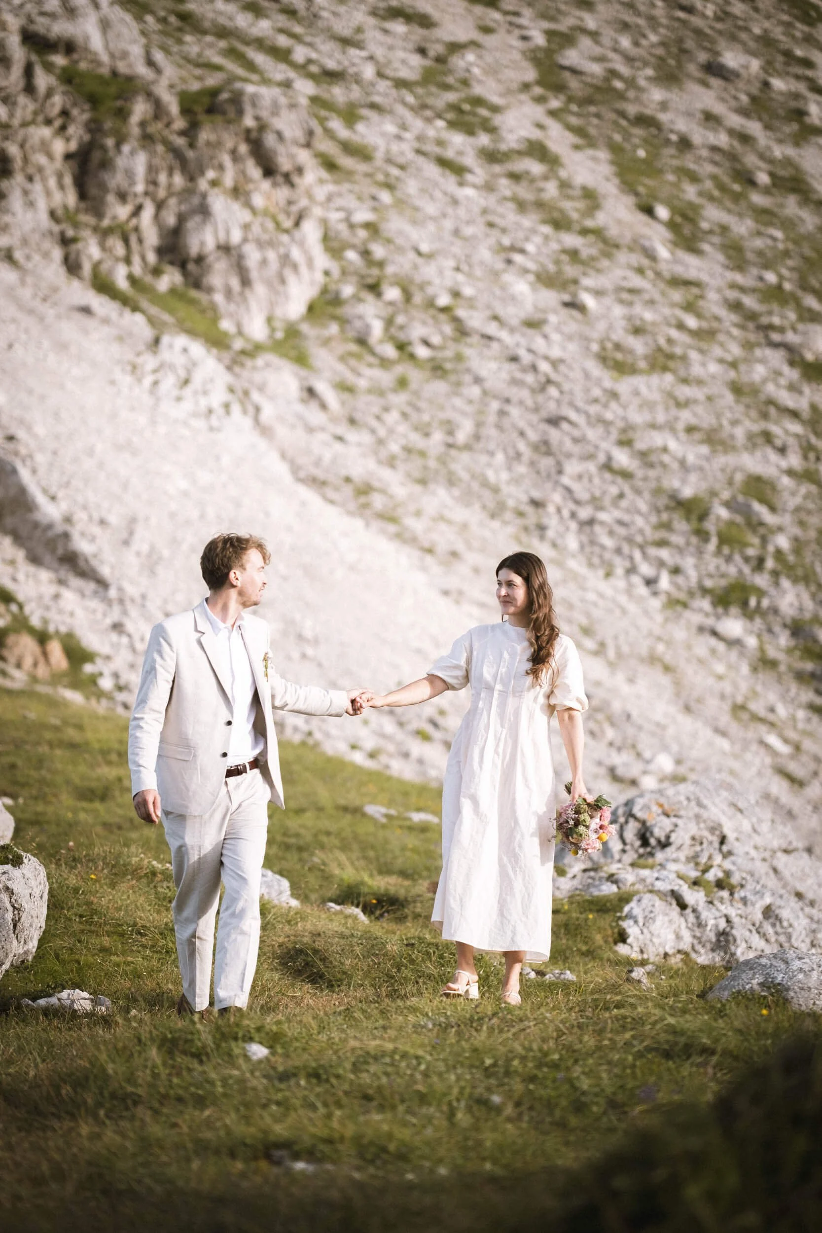 Ein Mann und eine Frau bei einer Hochzeit im Freien, umgeben von Steinen und Gräsern, die Frau hält einen Blumenstrauß, beide tragen leichte Sommerkleidung.