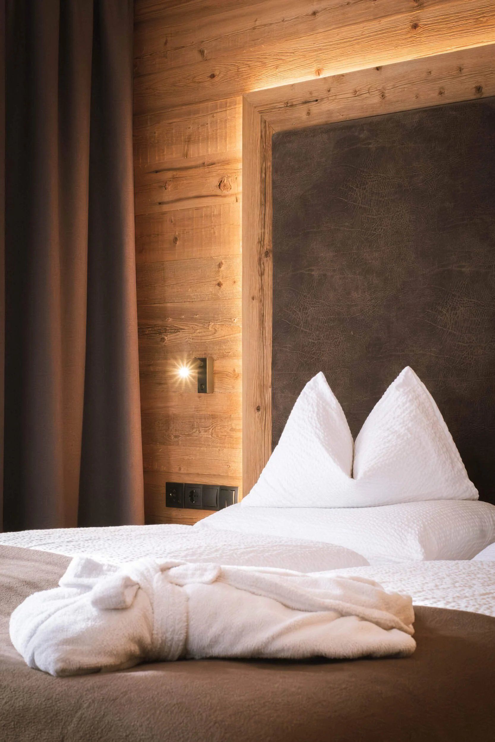 Ein Bett im Holzhausstil, mit weißen Kissen und einer Decke, vor einem Holzkopfkissen, seitliche Steckdosen und eine kleine Leselicht an der Wand.