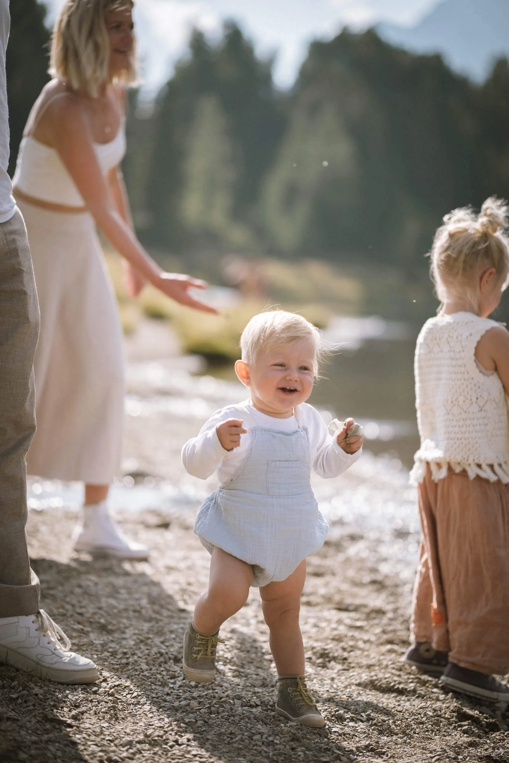 Ein glückliches Baby in Overalls läuft an einem Strand, während Erwachsene im Hintergrund stehen und lachen.