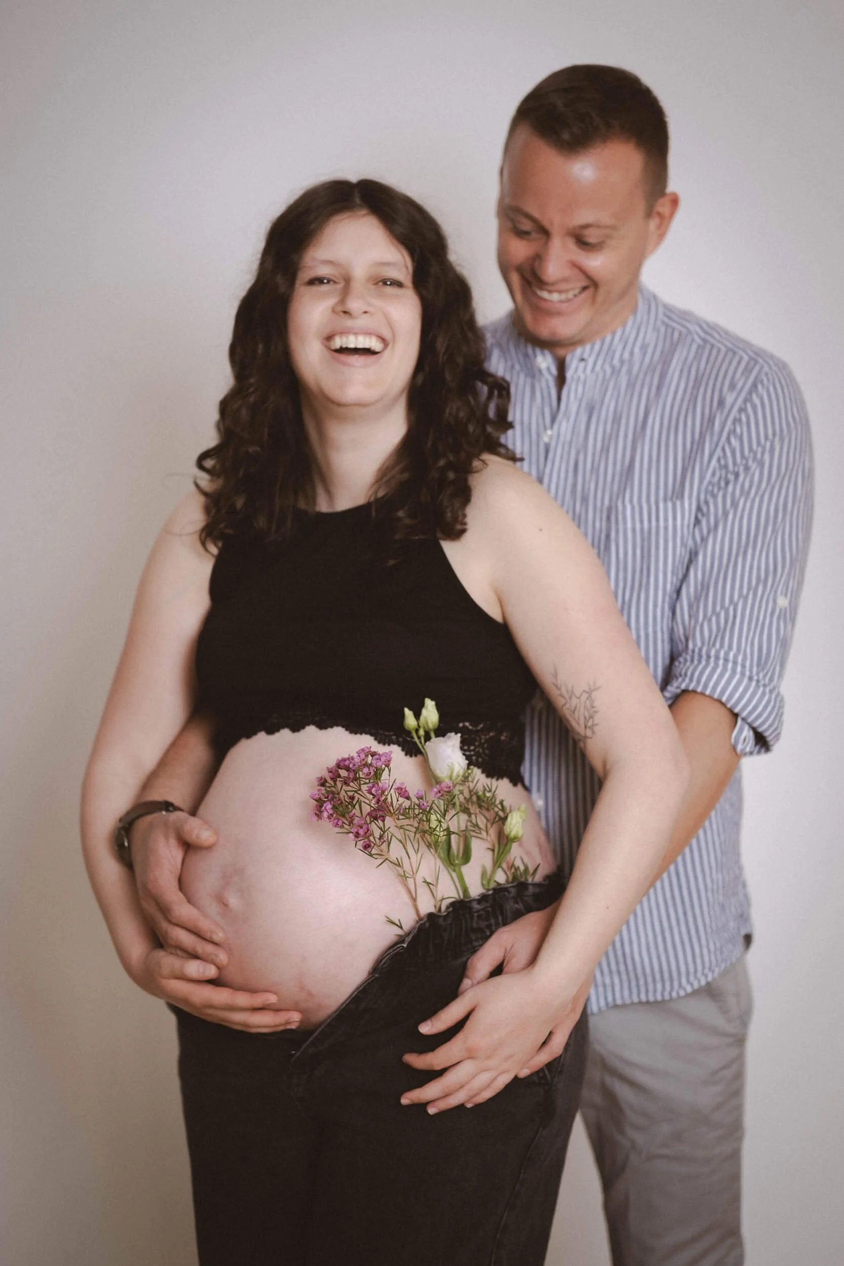 Ein lächelndes schwangeren Paar vor neutralem Hintergrund, die Frau hält Blumen vor ihren Bauch.