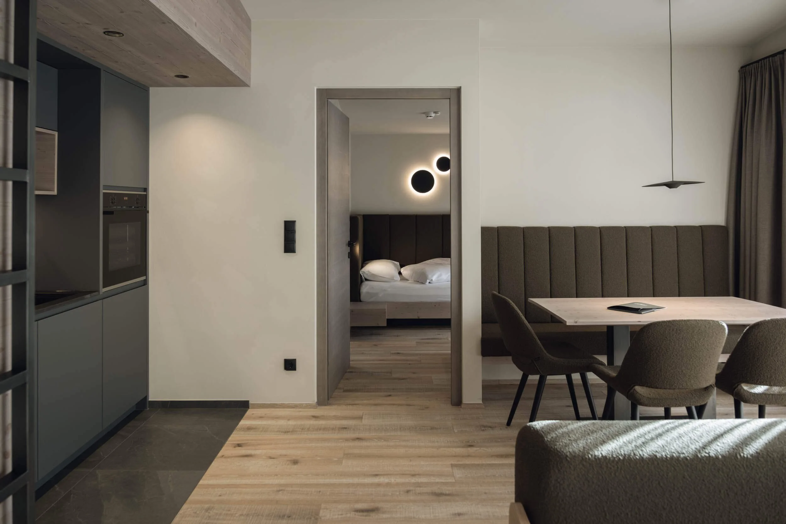 Ein modernes, minimalistisch eingerichtetes Zimmer mit Holzfußboden, einem Esstisch mit Stühlen, einer abgeschirmten Sitzecke, einer Küche und einem Schlafzimmer im Hintergrund.