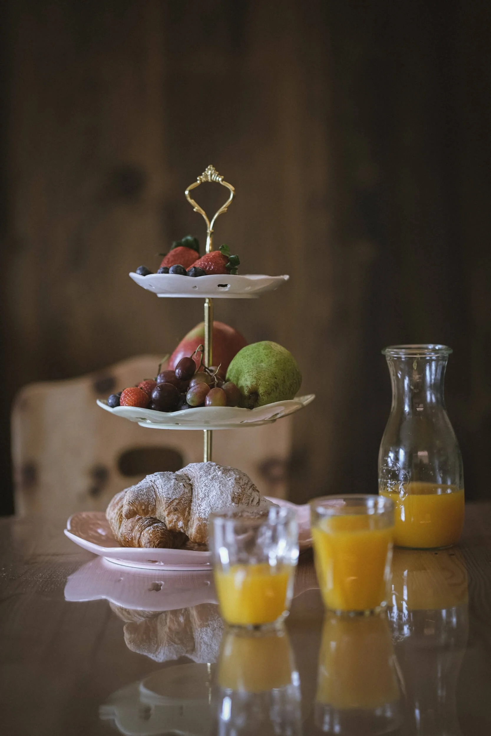 Ein Frühstückstisch mit einem dreistöckigen Obsttablett, Croissants, frisch gepresstem Orangensaft in einer Karaffe und Gläsern auf einem Holztisch.