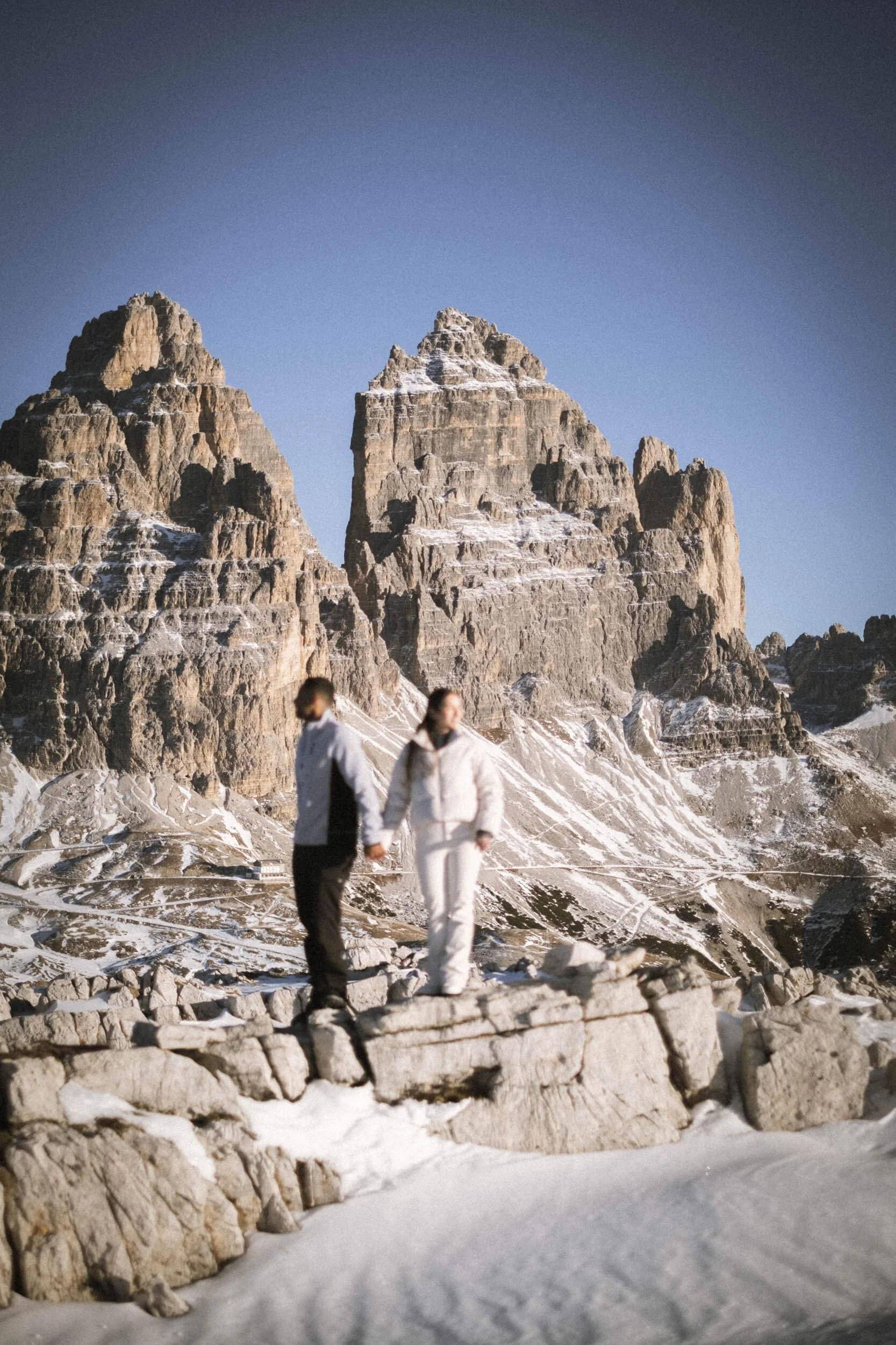 Ein Pärchen hält sich an den Händen, steht auf einem felsigen, verschneiten Gebiet vor einer beeindruckenden Bergkulisse bei klarem Himmel.