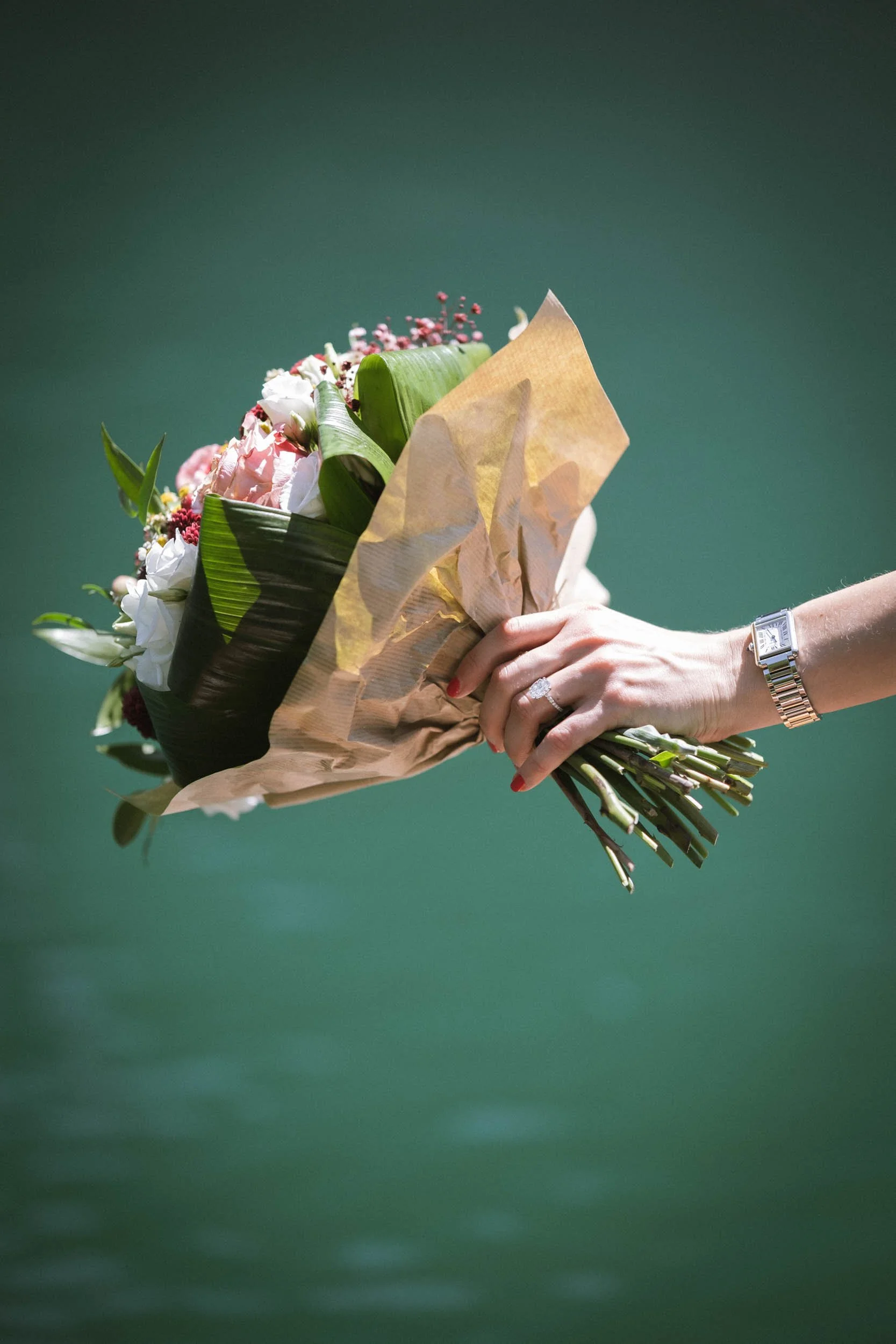 Eine Hand hält einen Blumenstrauß mit weißen, rosa und roten Blumen, eingewickelt in braunes Papier, vor grünem Hintergrund. Die Hand trägt eine silberne Armbanduhr und einen Ehering.