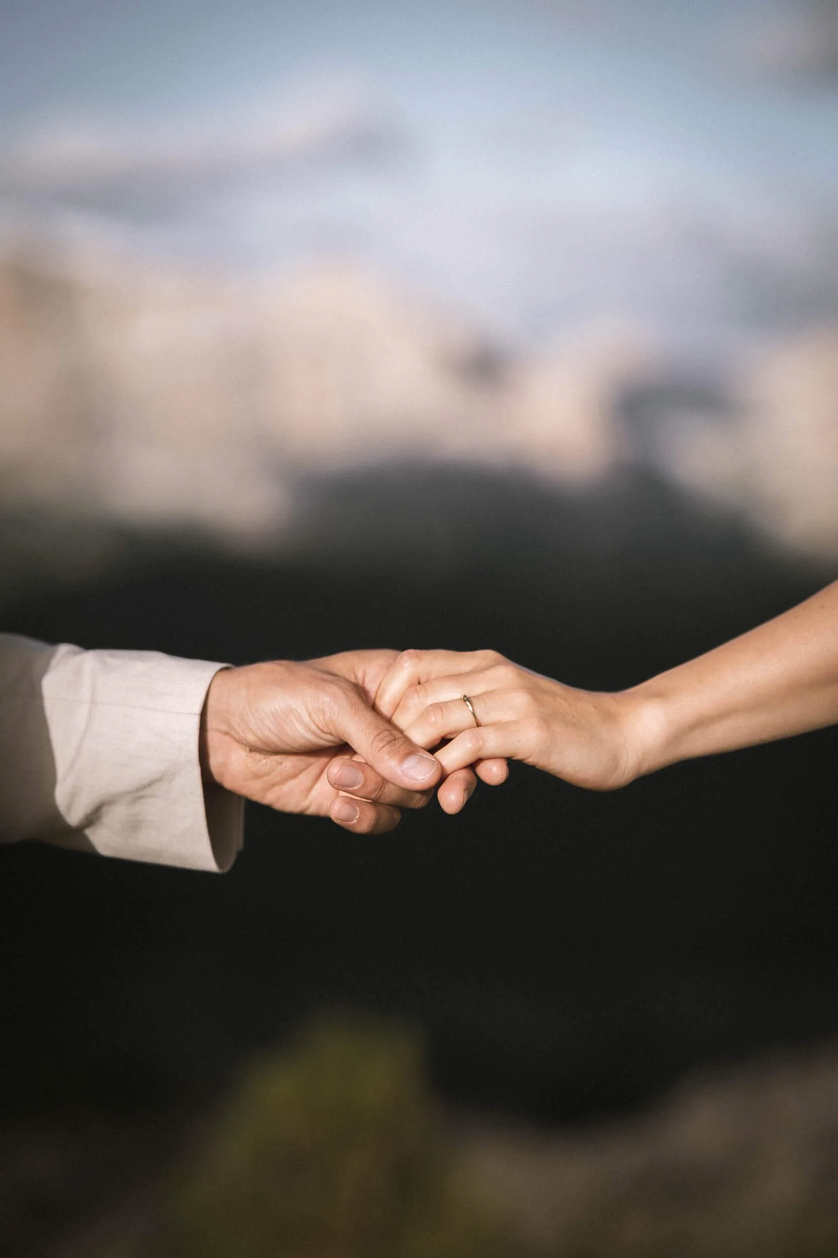 Zwei Hände, eine ältere Person und eine jüngere Person, halten sich im Hand. Der Hintergrund ist unscharf mit Himmel und Gebäude.