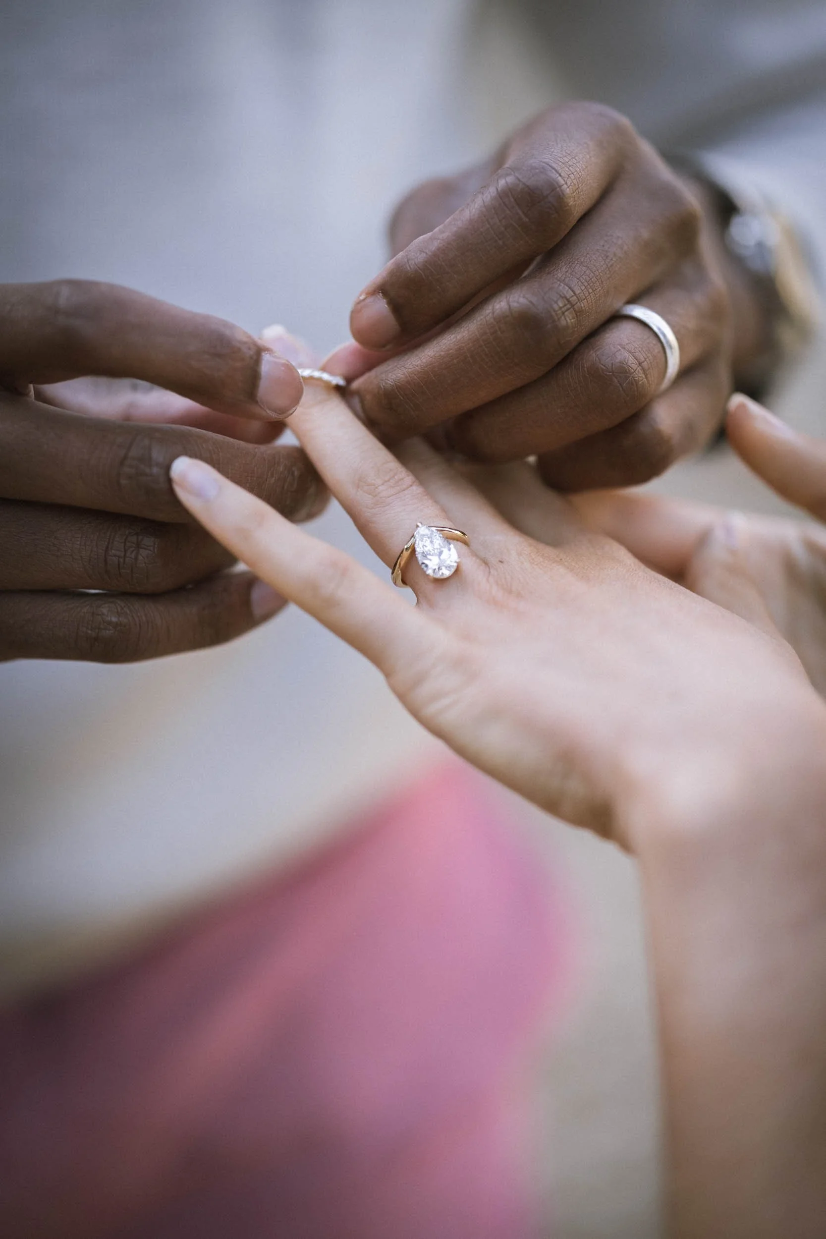 Zwei Hände, eine dunkle und eine helle, halten eine Hand mit einem großen Diamantsolitär-Ring. Es erscheint eine Verlobung oder Hochzeit.