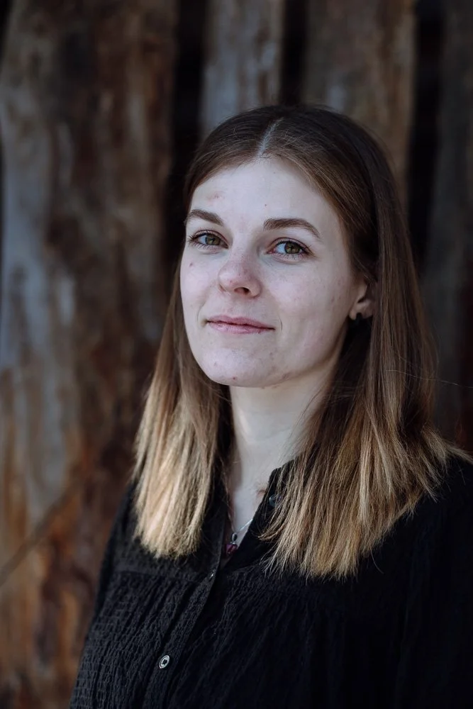 Ein junges Mädchen mit braunem Haar und blauen Augen vor einem Baumstamm, trägt eine schwarze Jacke.