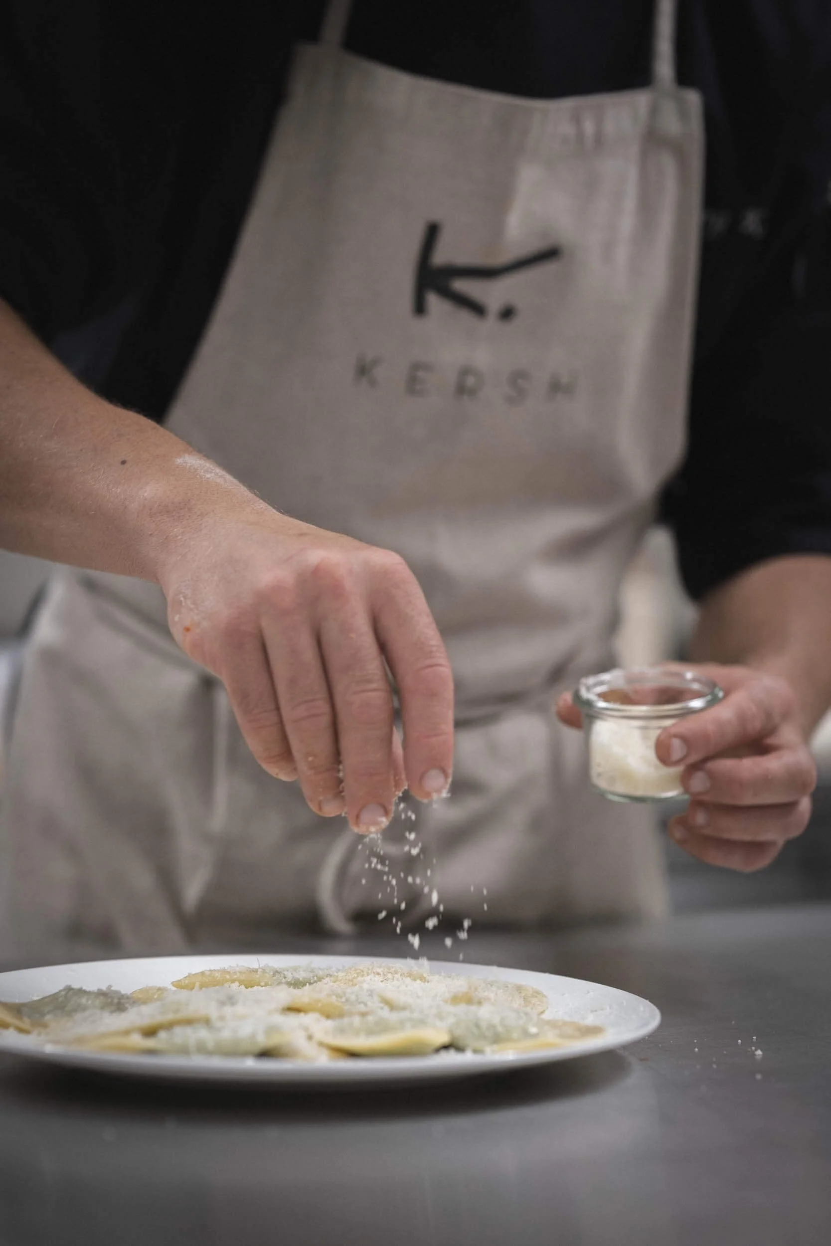 Person bestreut Ravioli mit Parmesan-Käse in einer Küche.