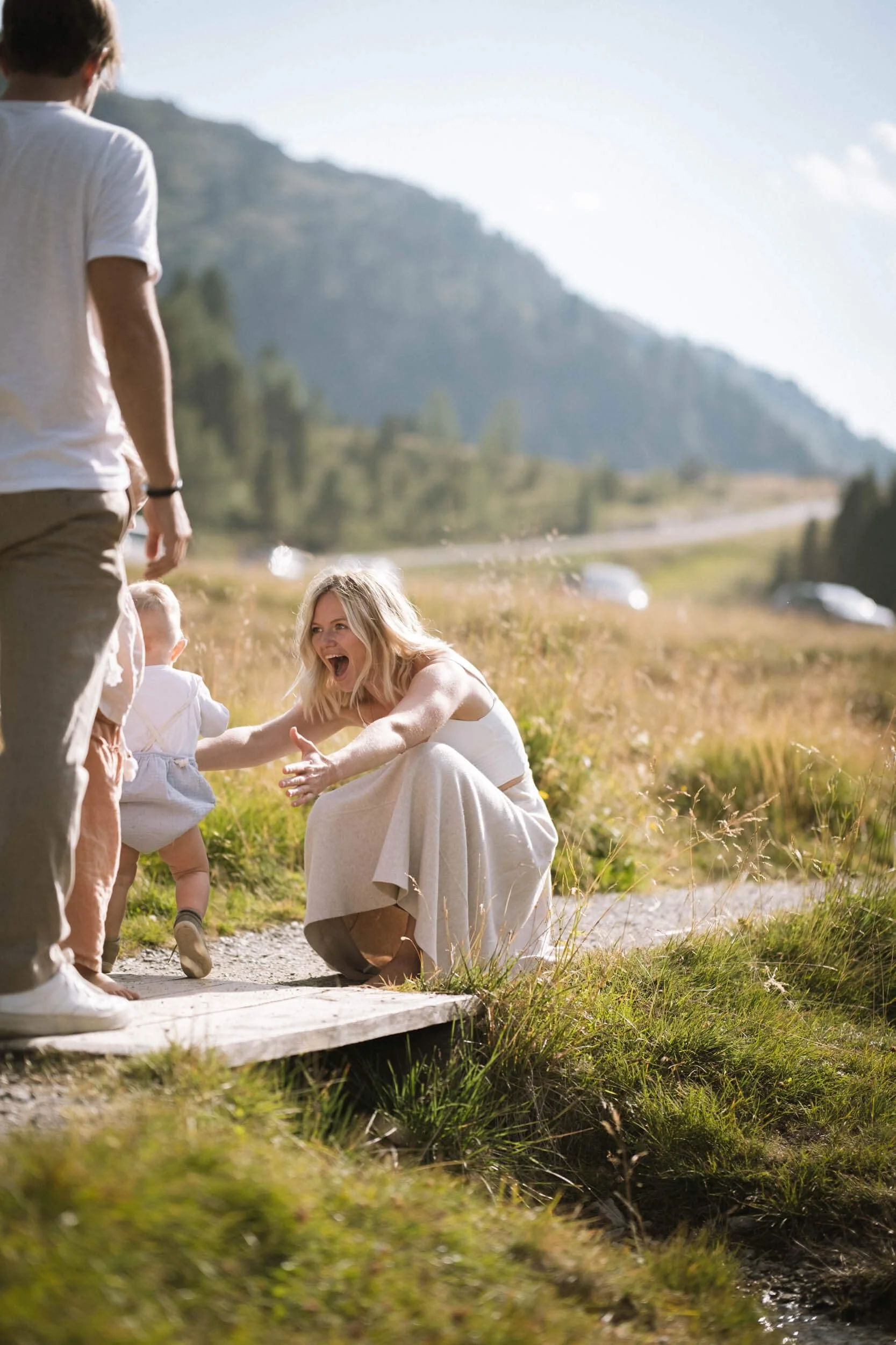 Frau im weißen Kleid freut sich, während sie ein Kind auf einem Holzsteg im Freien begrüßt, im Hintergrund Berge, Wiese und Himmel.