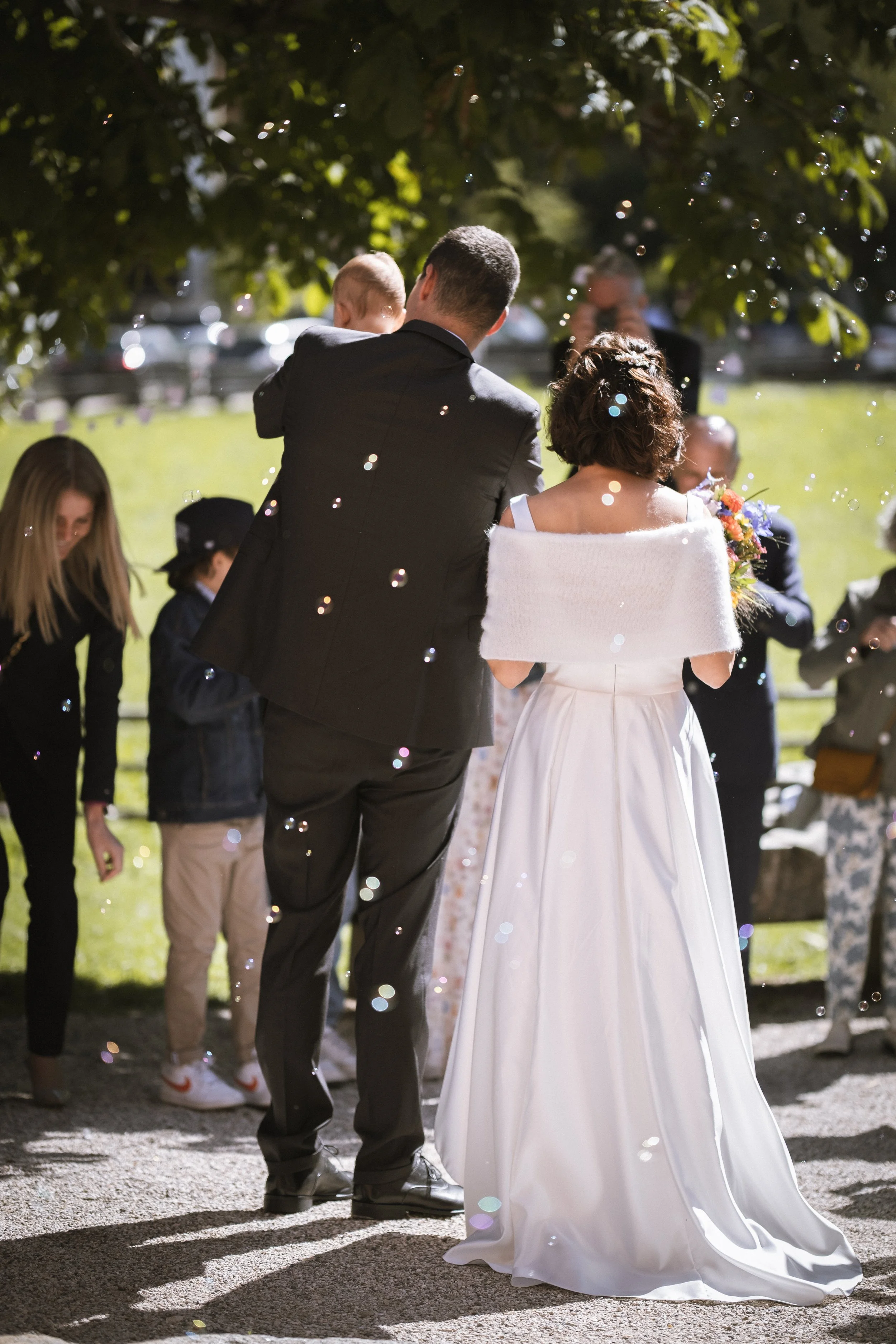 Hochzeitspaar steht im Freien, umgeben von Gästen, bei Sonnenschein, mit Seifenblasen in der Luft.