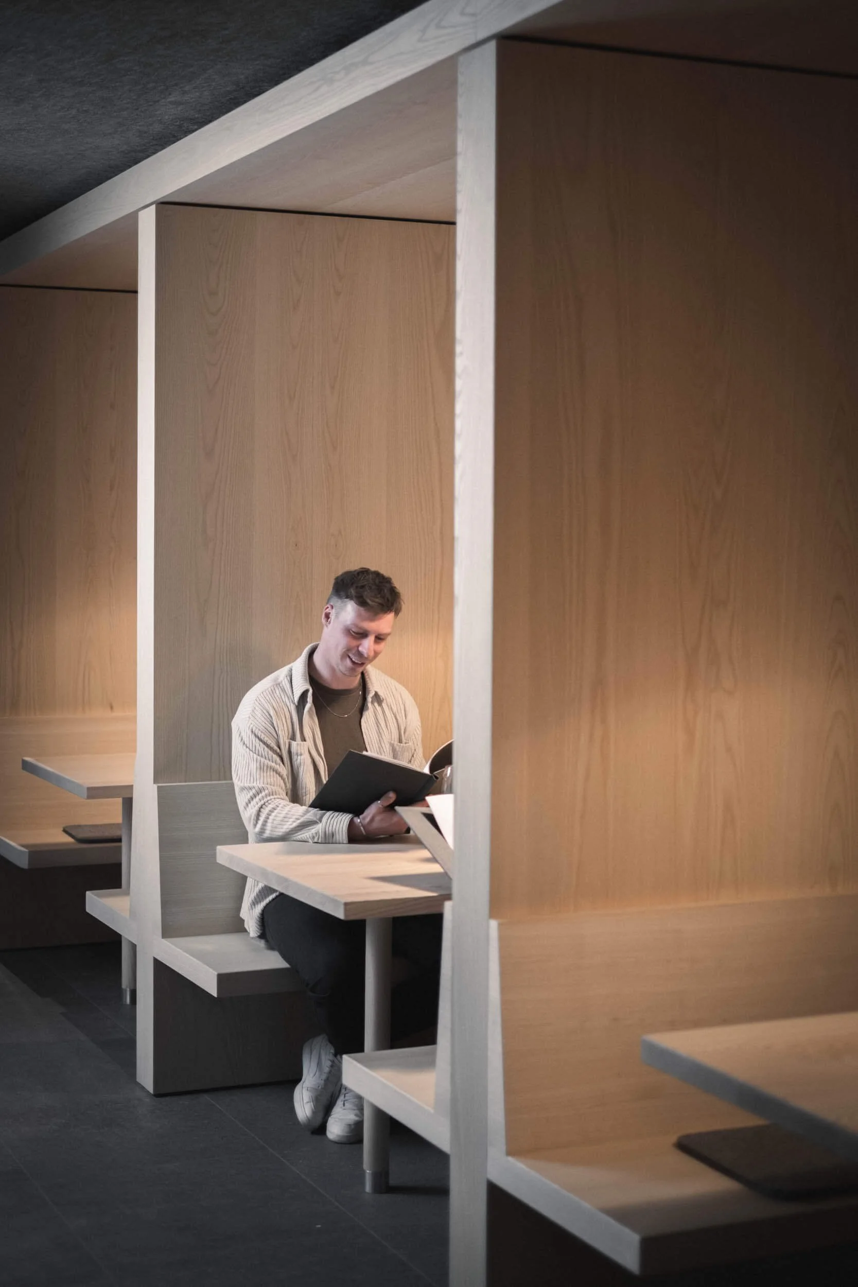 Ein junger Mann liest in einem Buch in einem modern gestalteten Pausenraum mit Holzschutzwänden und Sitznischen.