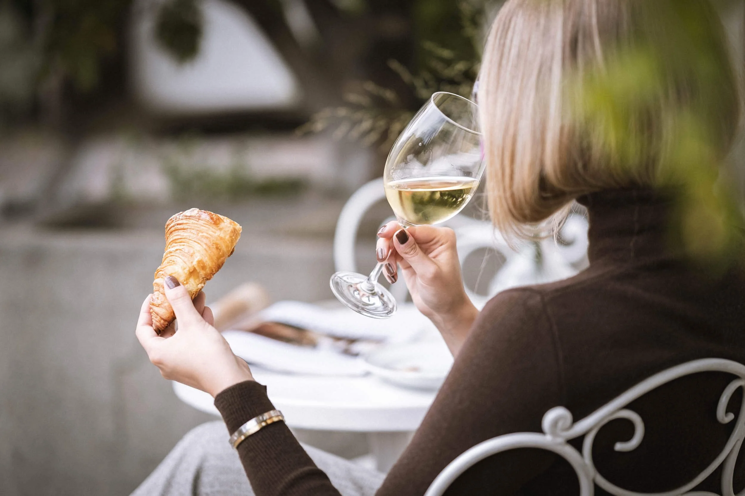 Eine Frau sitzt im Freien, hält ein Glas Weißwein in der einen Hand und ein Croissant in der anderen, vor einem kleinen Tisch mit weiteren Gegenständen, während sie sich entspannt