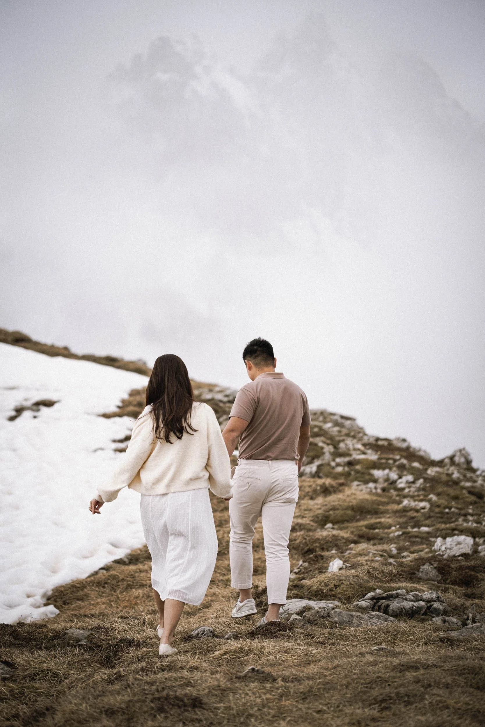 Ein Mann und eine Frau gehen auf einem Bergpfad, umgeben von Schnee und Fels, bei bewölktem Himmel.