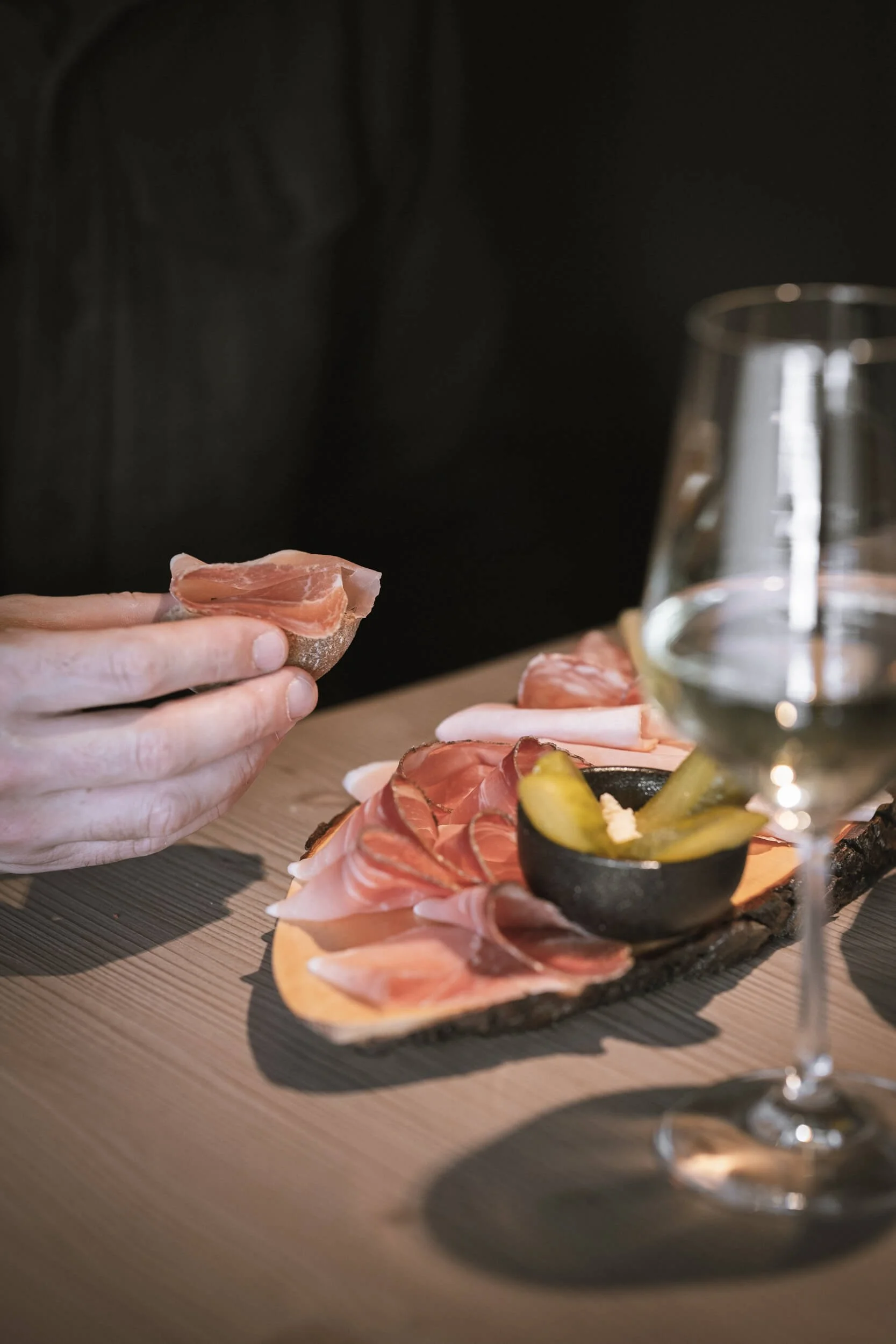 Nahaufnahme von einer Person, die eine Scheibe Schinken hält, mit einer Auswahl an Schinken und anderen Wurstsorten auf einem Holzbrett, einem kleinen Schwarzglas mit Senf, einem Weinglas und einem Wasserkrug auf einem Tisch.