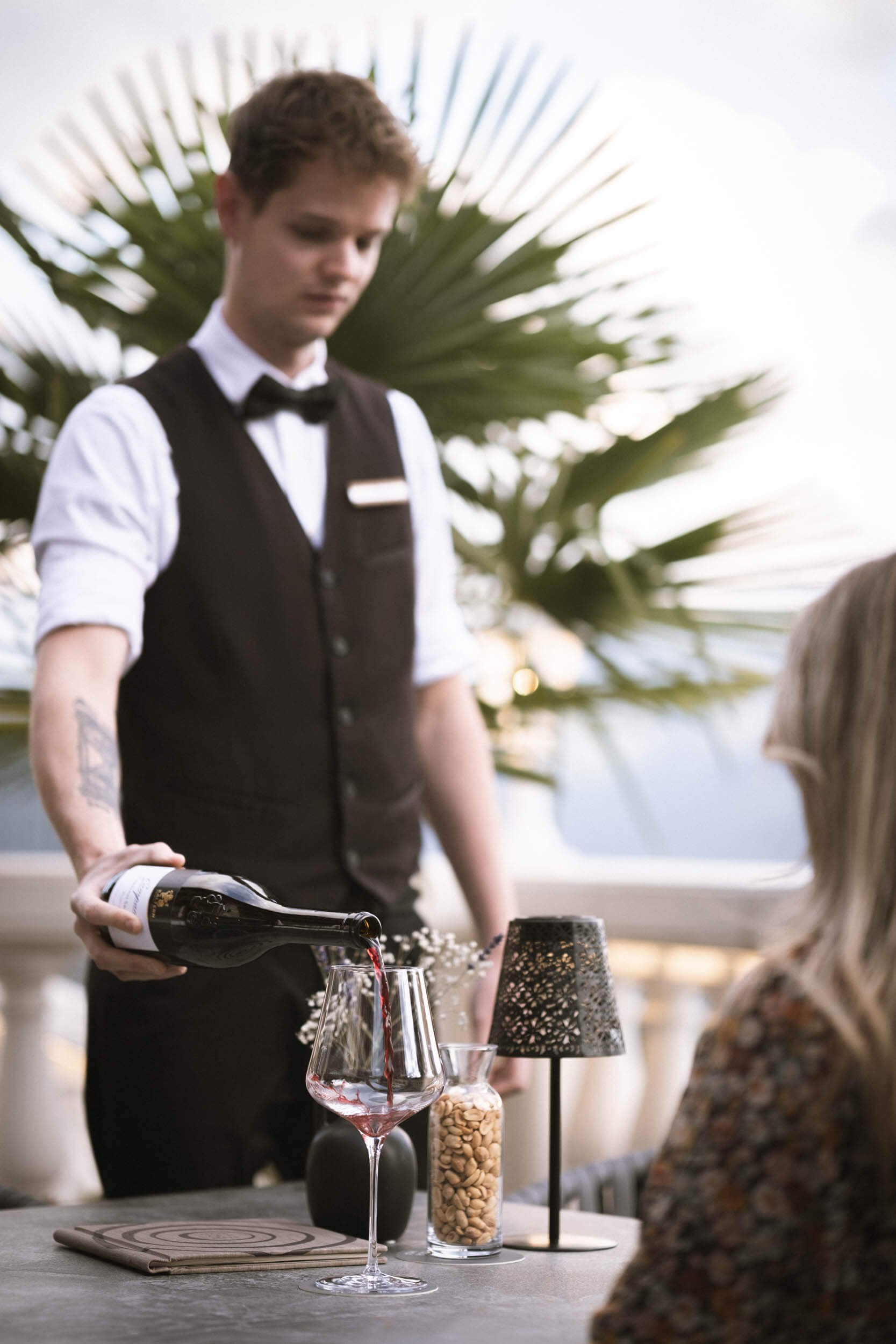 Ein junger Mann in Schwarz-Weiß-Gastronomieuniform serviert im Freien Wein an eine Frau.