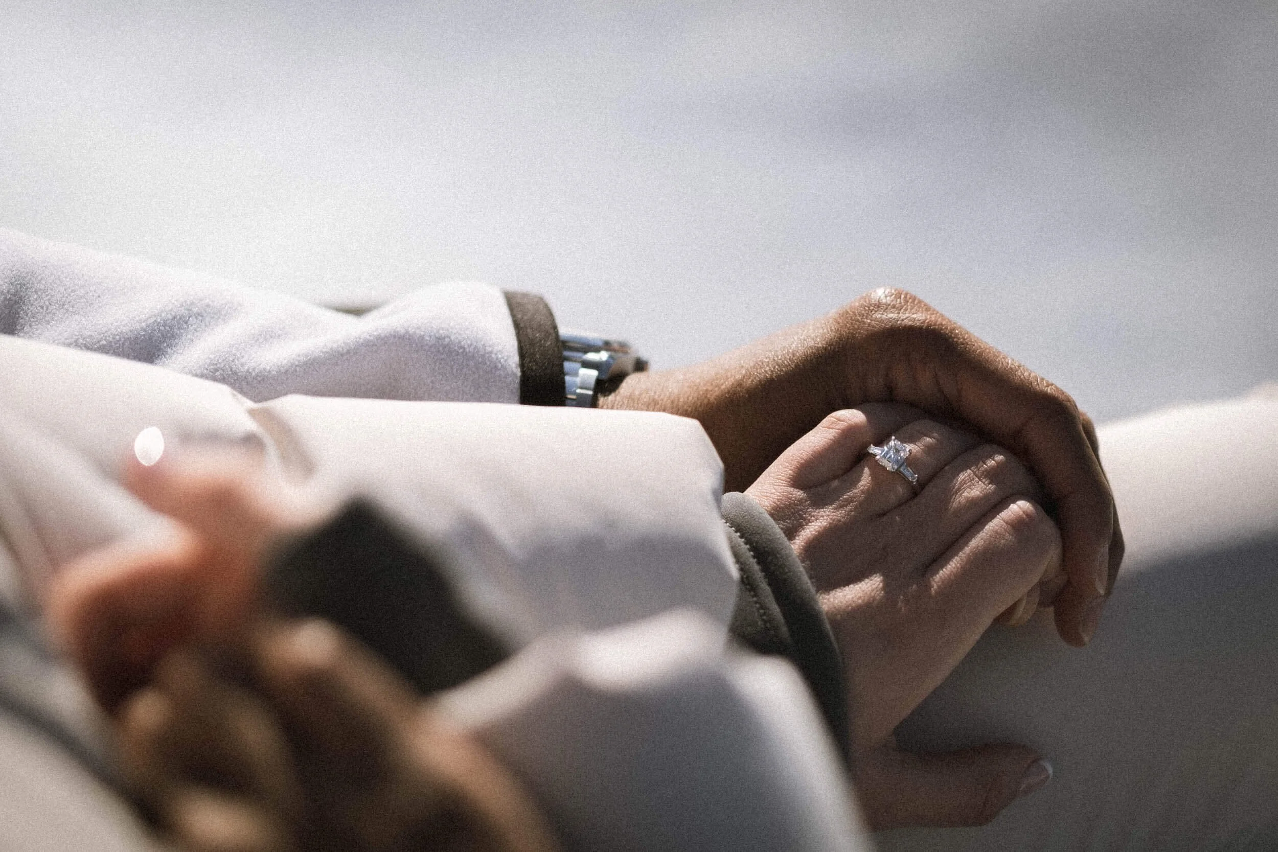 Nahaufnahme von zwei Händen, die sich halten, eine mit einem Diamant-Ehehandring, die andere mit einem dunkleren Hautton, im Freien bei Tageslicht.