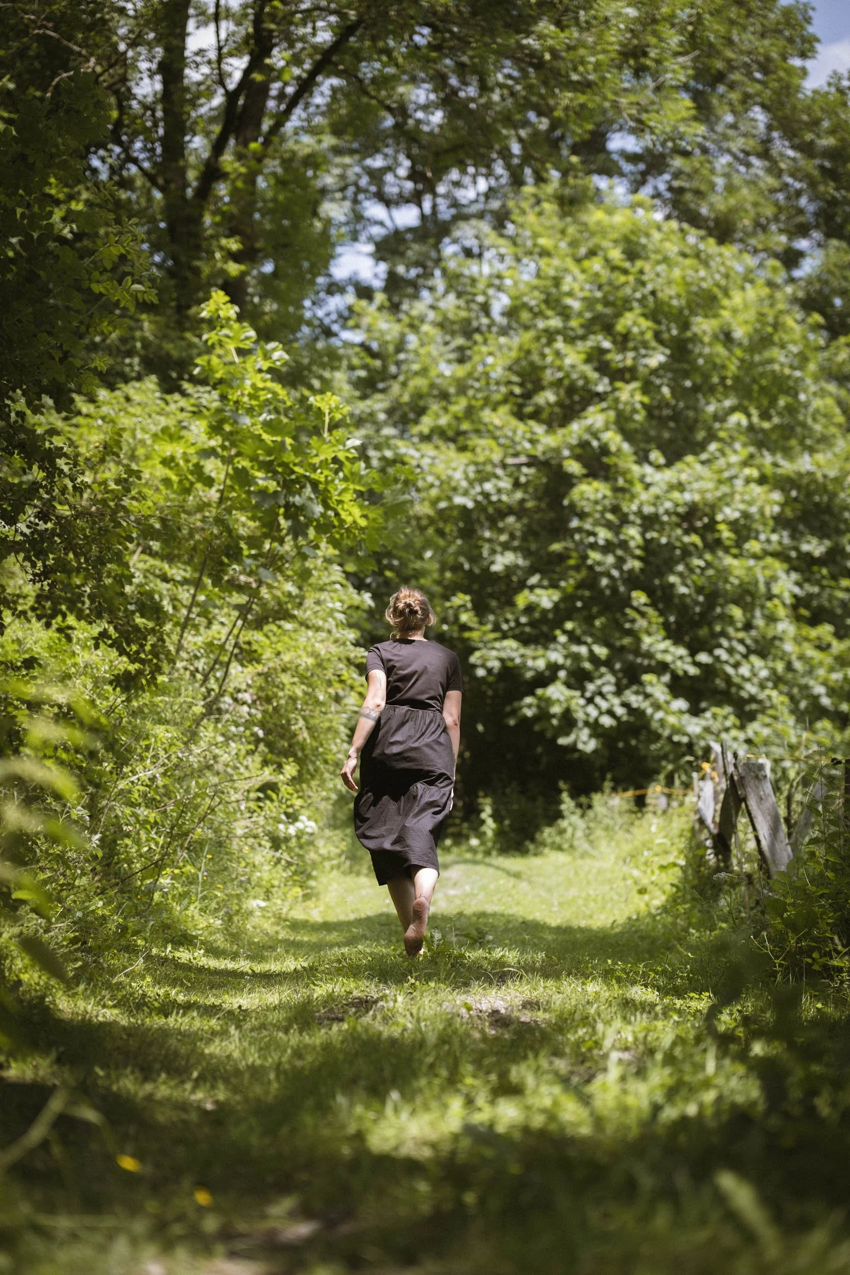 Eine Frau läuft barfuß auf einem grasbewachsenen Weg in einem dichten grünen Wald, gesehen von hinten.