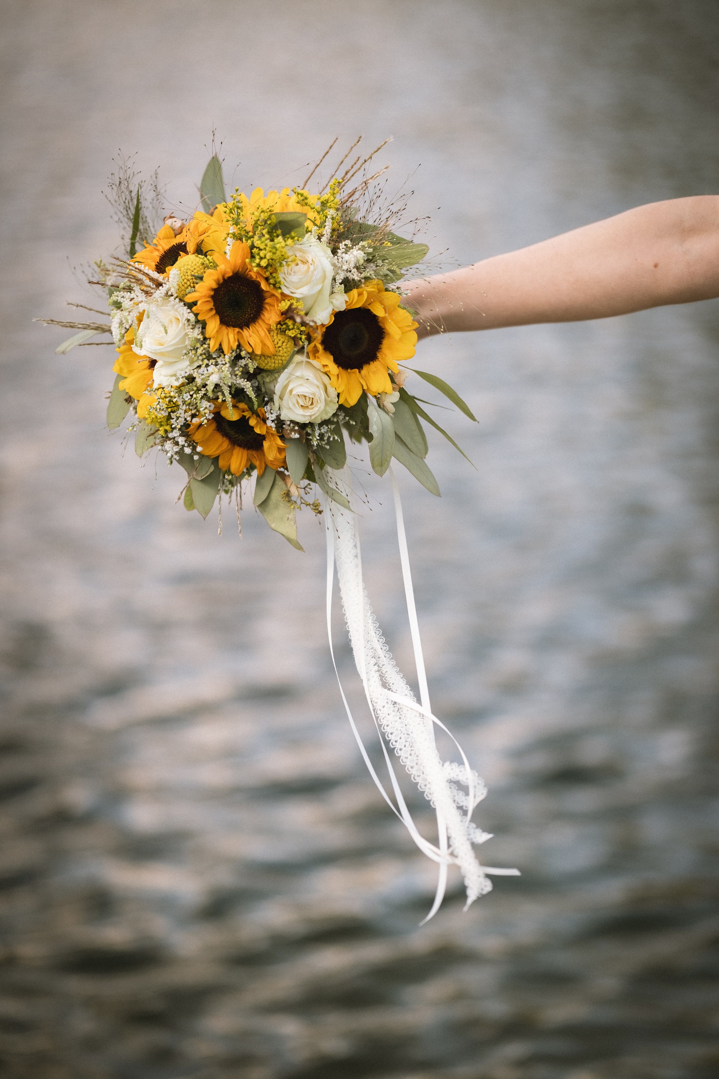 Hand hält einen Brautstrauß mit Sonnenblumen, weißen Rosen, Gelbblumen und grünem Laub, vor Wasser im Hintergrund.
