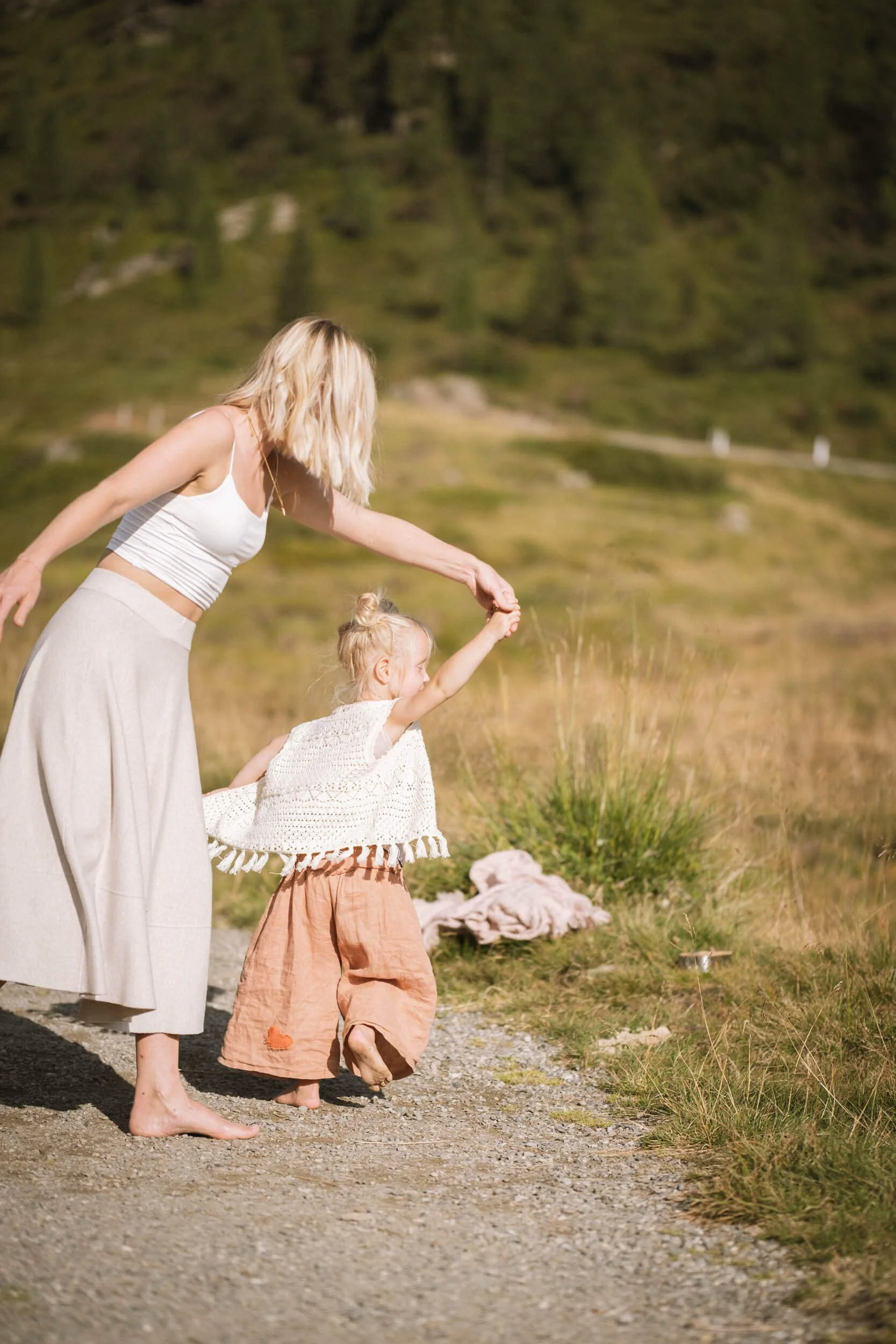 Eine Frau und ein kleines Mädchen spielen barfuß auf einem Weg im Freien, mit einer grünen, bergigen Landschaft im Hintergrund. Das Mädchen wirft etwas in die Luft, während die Frau leicht nach vorn gebeugt ist und sie dabei beobachtet.
