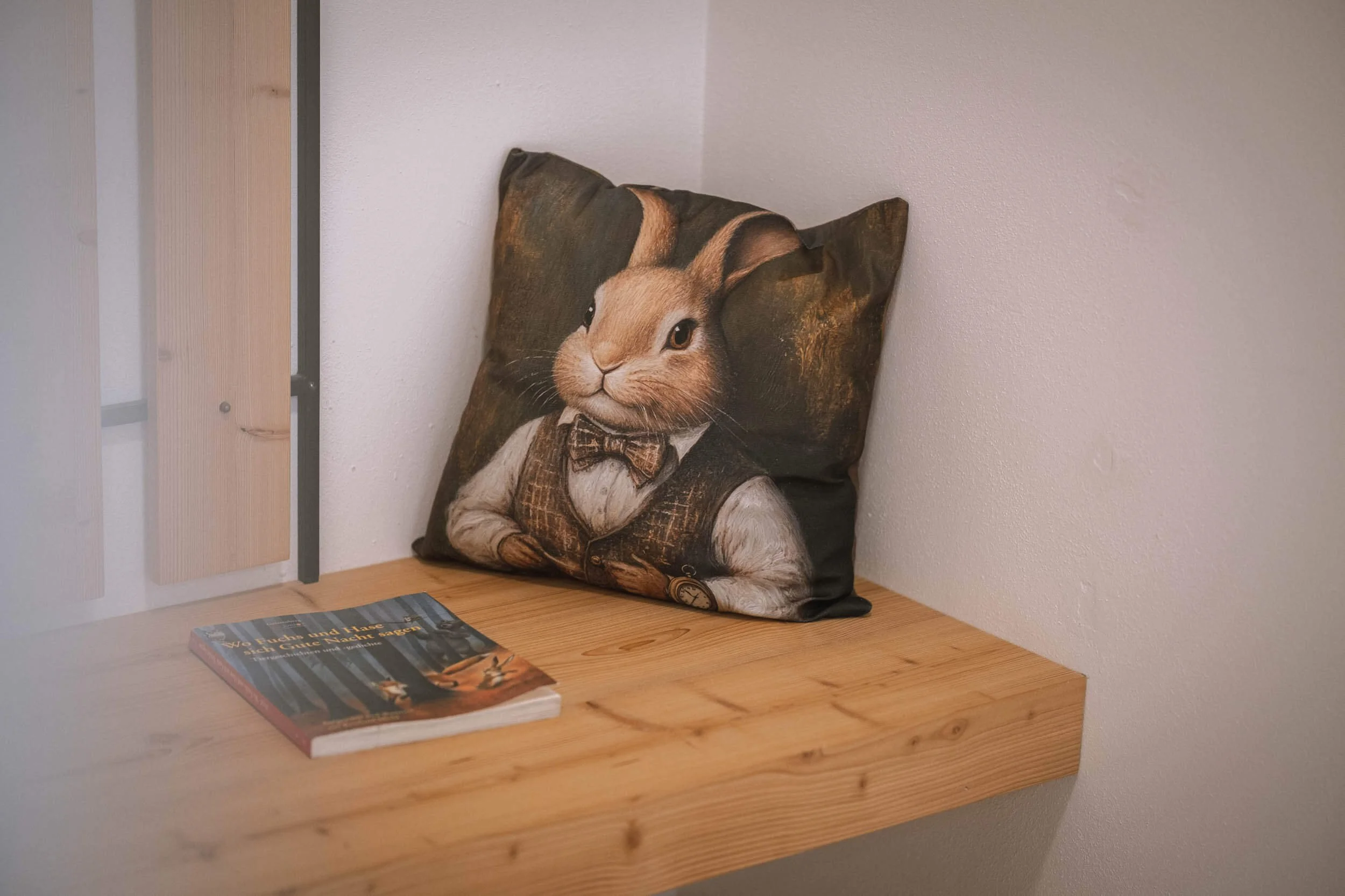 Ein Kissen mit einem Hasengesicht in einem Anzug und Fliege, auf einem Holzregal neben einem Buch.