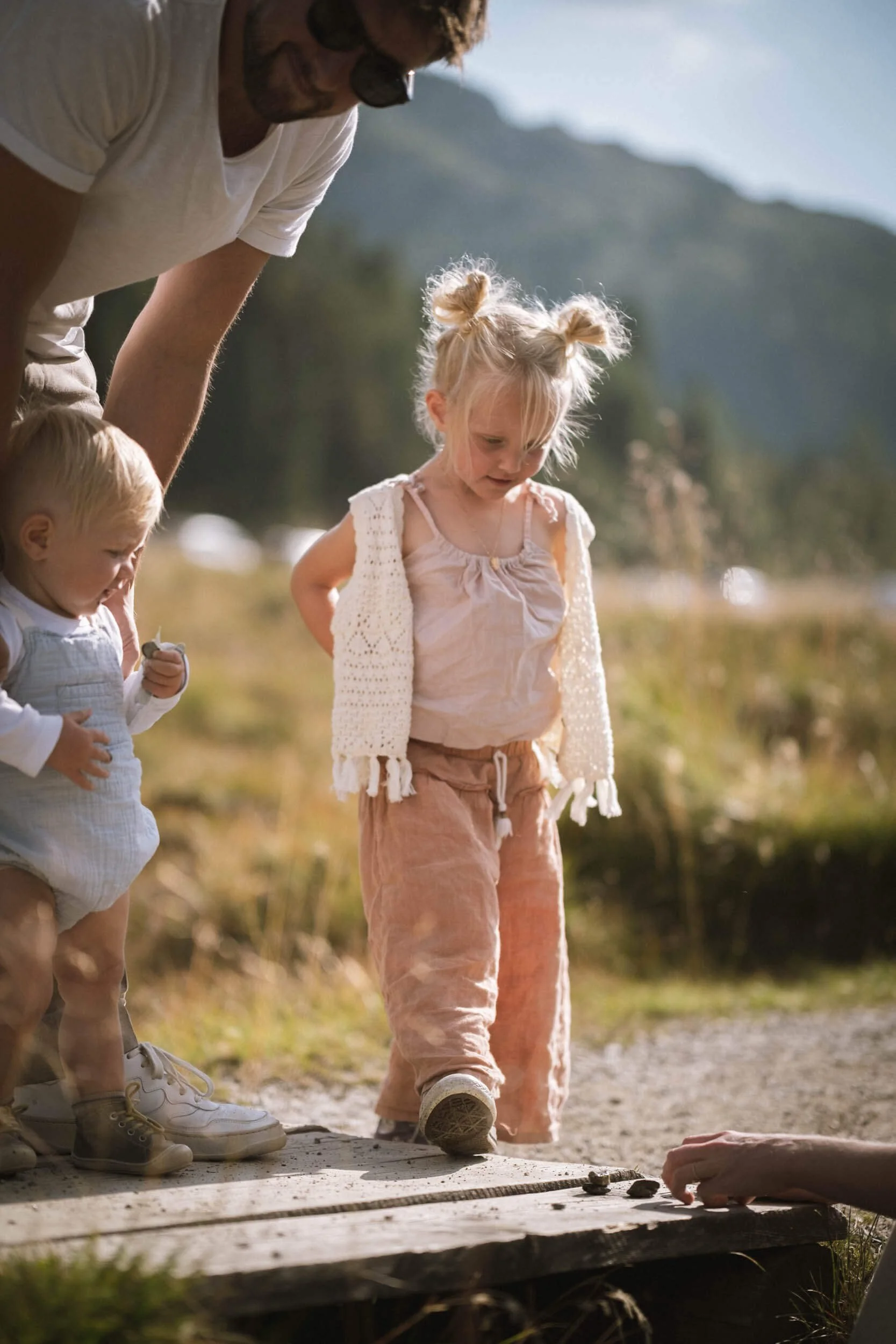 Zwei kleine Kinder, ein Junge und ein Mädchen, mit einem Erwachsenen im Freien auf einer Holzplattform. Das Mädchen trägt ein rosa Outfit und eine weiße Strickjacke, der Junge trägt eine weiße Latzhose, und der Erwachsene trägt Sonnenbrille. Sie sche