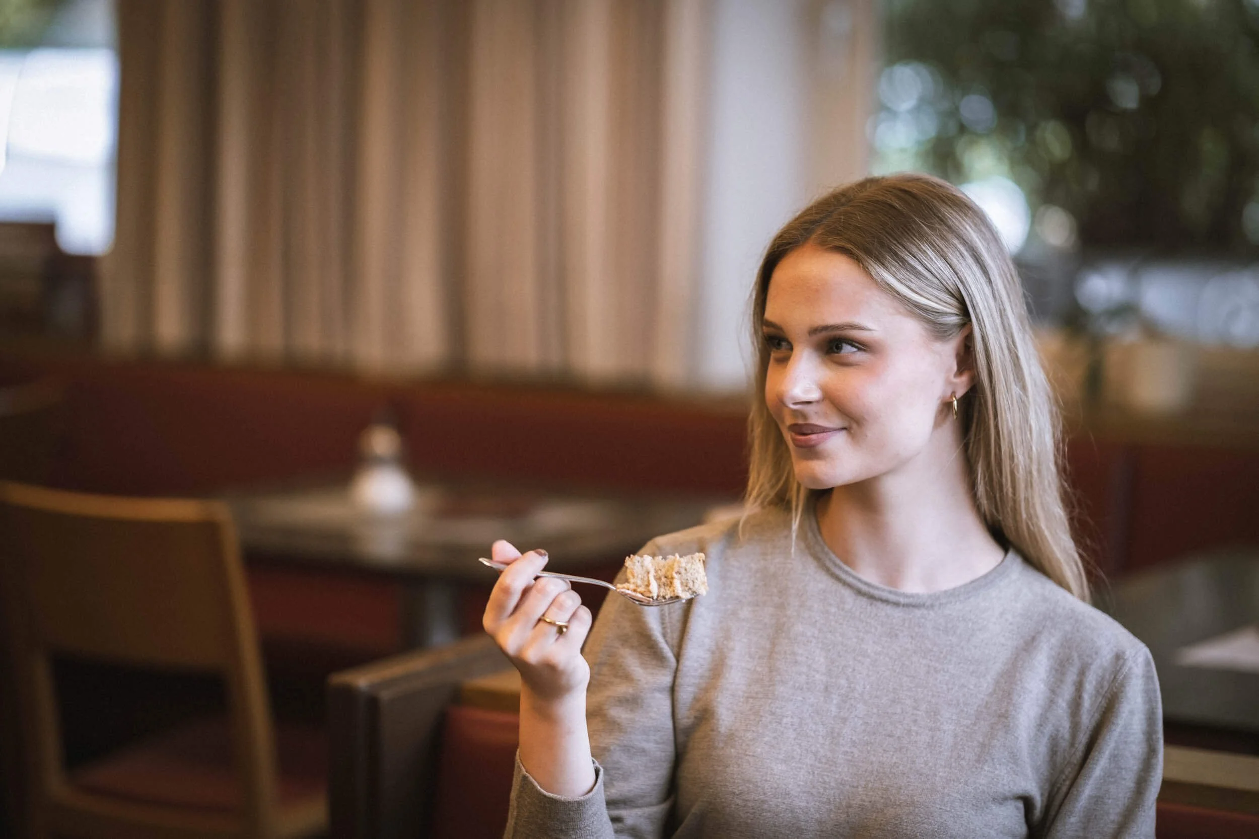 Junge Frau mit langen, blonden Haaren und grauem Oberteil sitzt in einem Restaurant und isst ein Stück Kuchen, während sie in die Kamera lächelt.