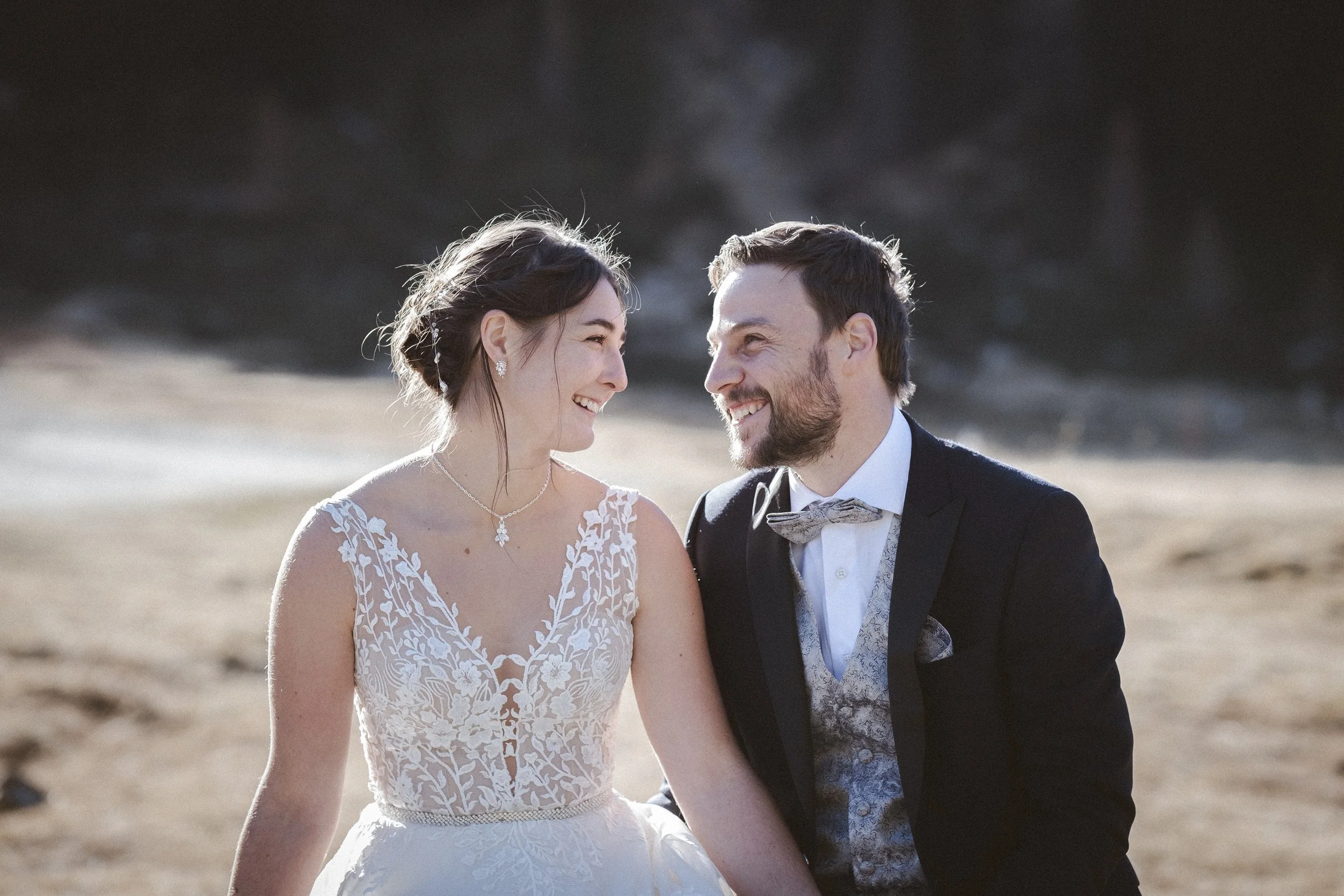 Ein glückliches Brautpaar sitzt zusammen im Freien bei Sonnenuntergang, die Frau trägt ein weißes Hochzeitskleid mit Spitzenmuster, der Mann trägt einen schwarzen Anzug mit einer Schleife. Beide lachen sich an, der Hintergrund ist natürlich und ruhig